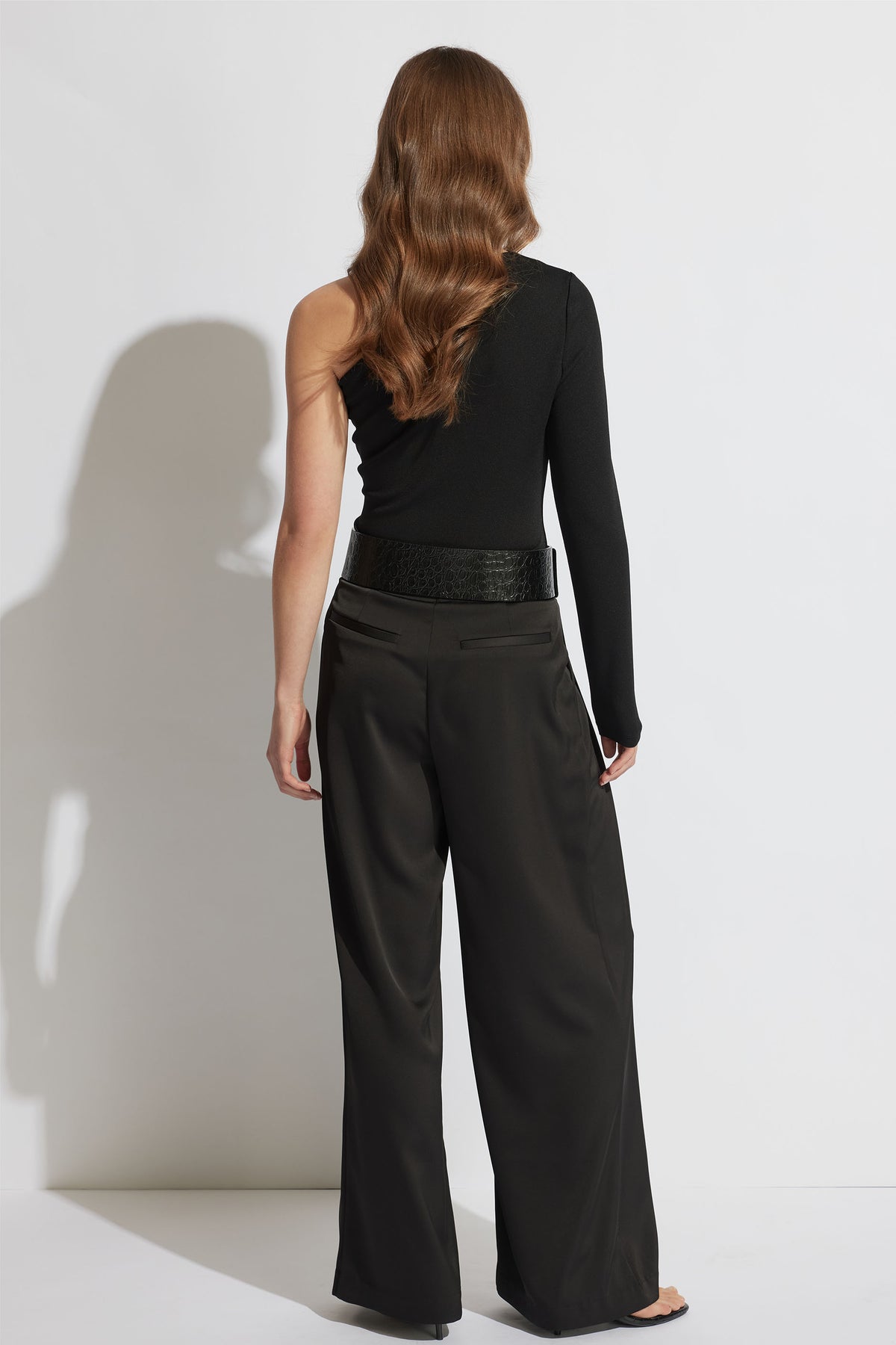 Woman wearing a black one-shoulder top with one long sleeve paired with wide-leg black trousers and a textured belt