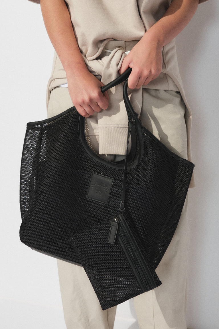 Person holding a black mesh structured tote bag with matching pouch, wearing beige casual pants and a tied sweatshirt.