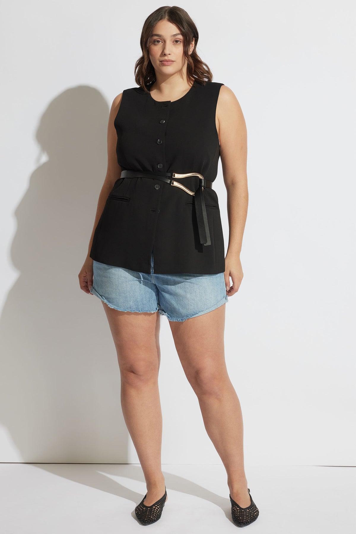 Woman wearing a sleeveless black buttoned top with a belt, paired with light blue rolled cuff denim shorts and black shoes