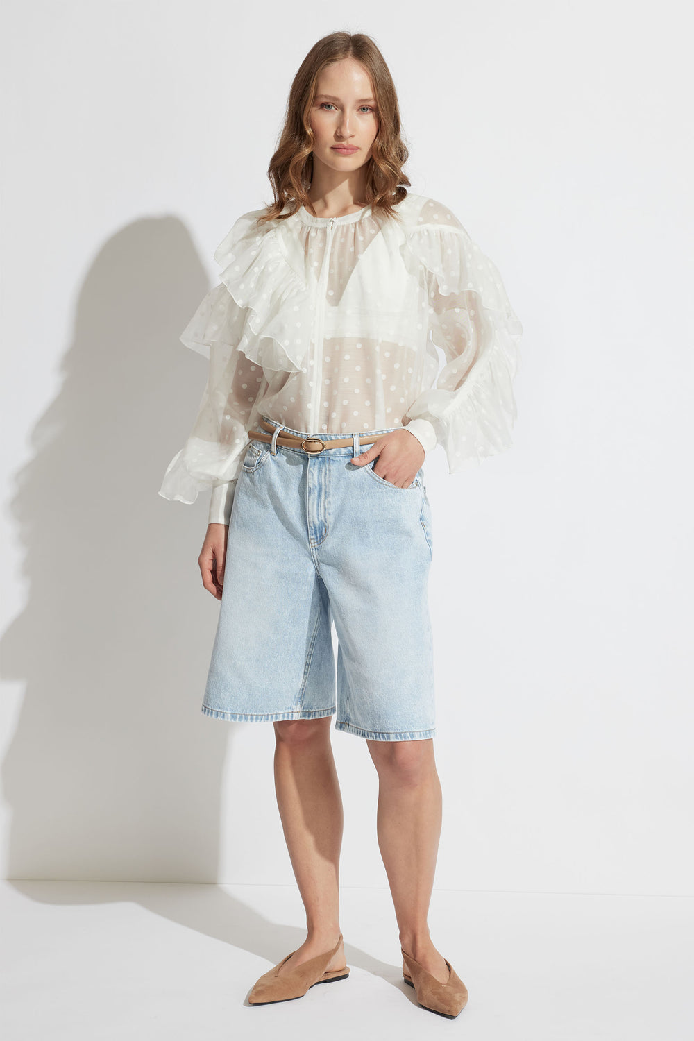 Woman wearing sheer white spot ruffle top paired with light-wash denim shorts and tan flats in natural light