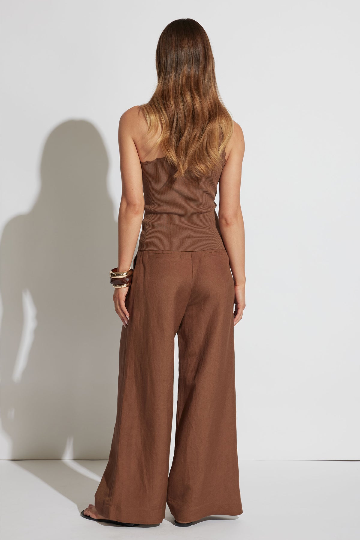 Woman wearing a brown one-shoulder tank paired with wide-leg pants and stacked bangles, shown from behind.