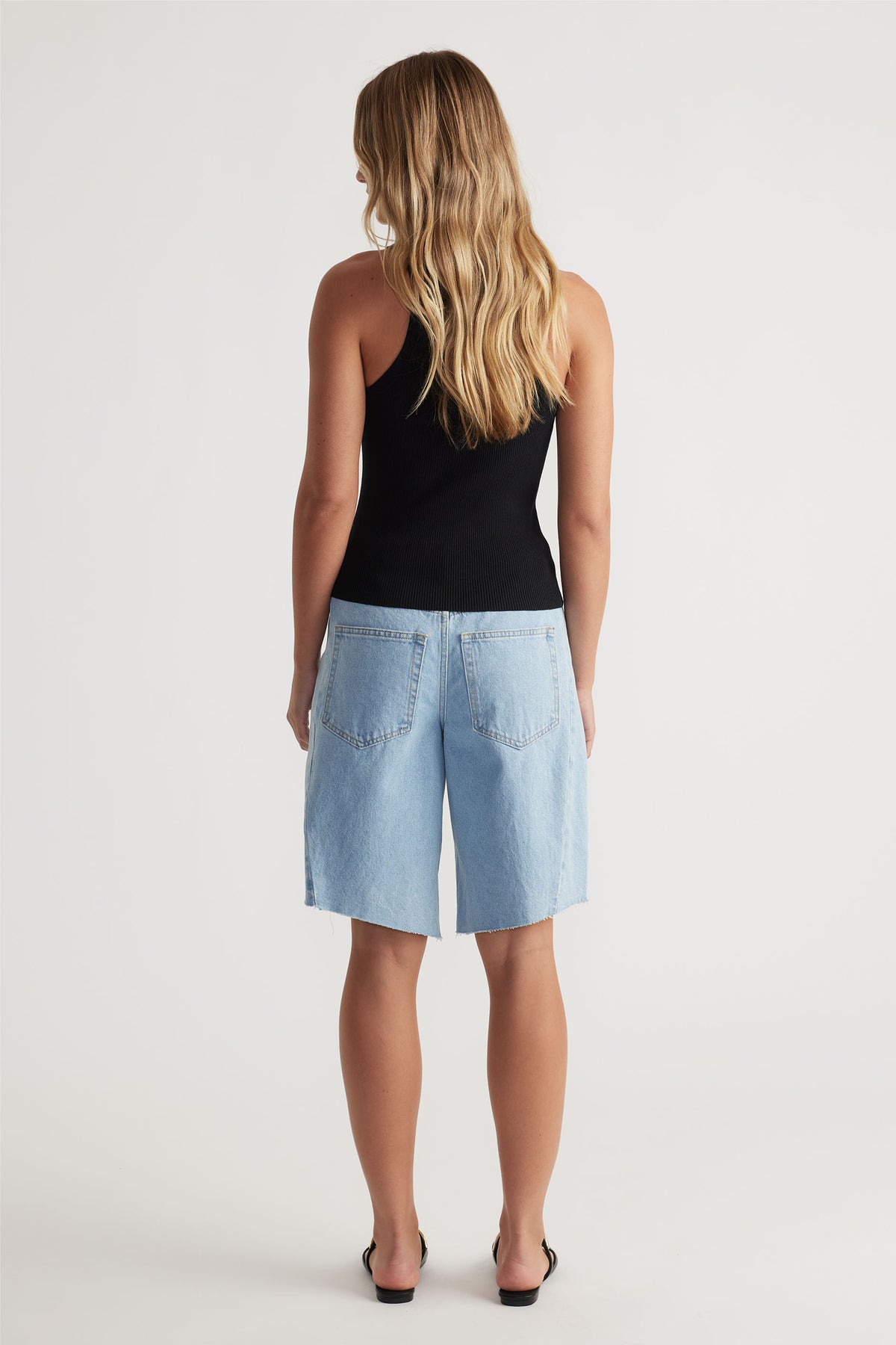 Back view of a woman wearing a black racer back knit tank paired with relaxed light-wash denim bermuda shorts.