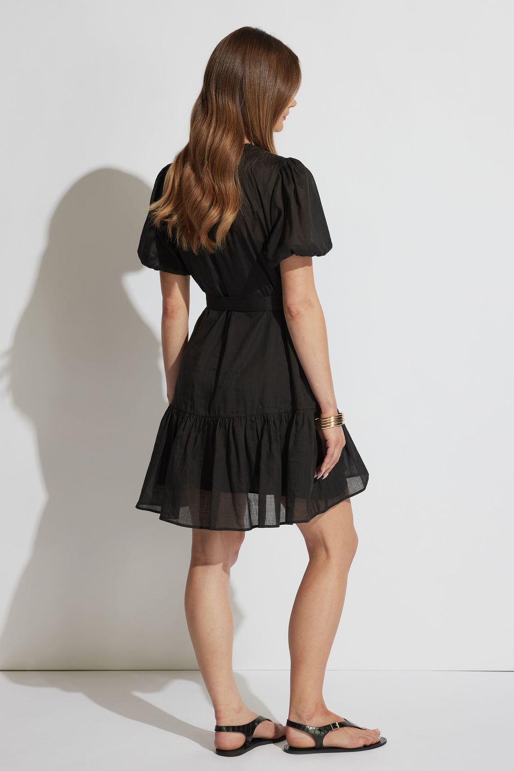 Woman pictured from behind wearing a black, short puff-sleeve dress with a gathered skirt and black sandals