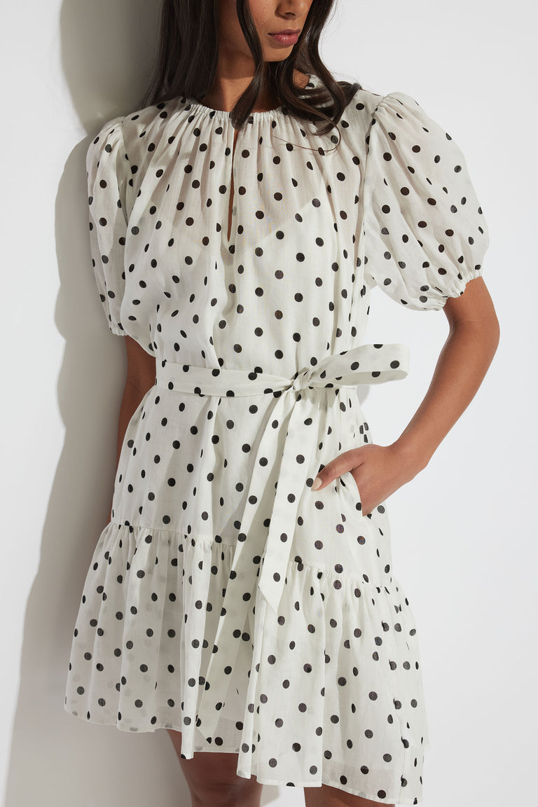 Woman wearing a white mini dress with black polka dots, puff sleeves, a gathered neckline, and a waist tie belt.