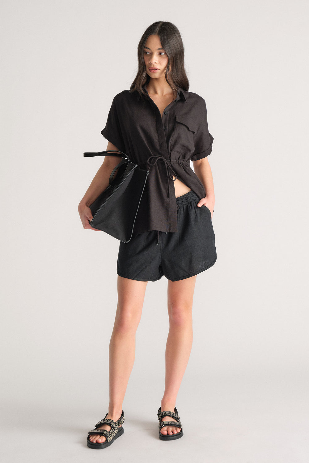 Woman wearing a black linen blend drawstring shirt with relaxed black shorts, carrying a large black tote bag.