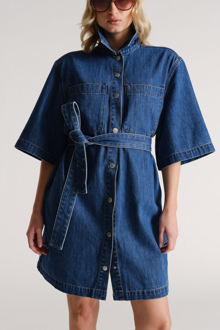 Woman wearing a dark blue denim short dress with wide sleeves, front buttons, chest pockets, and a tied waist belt.