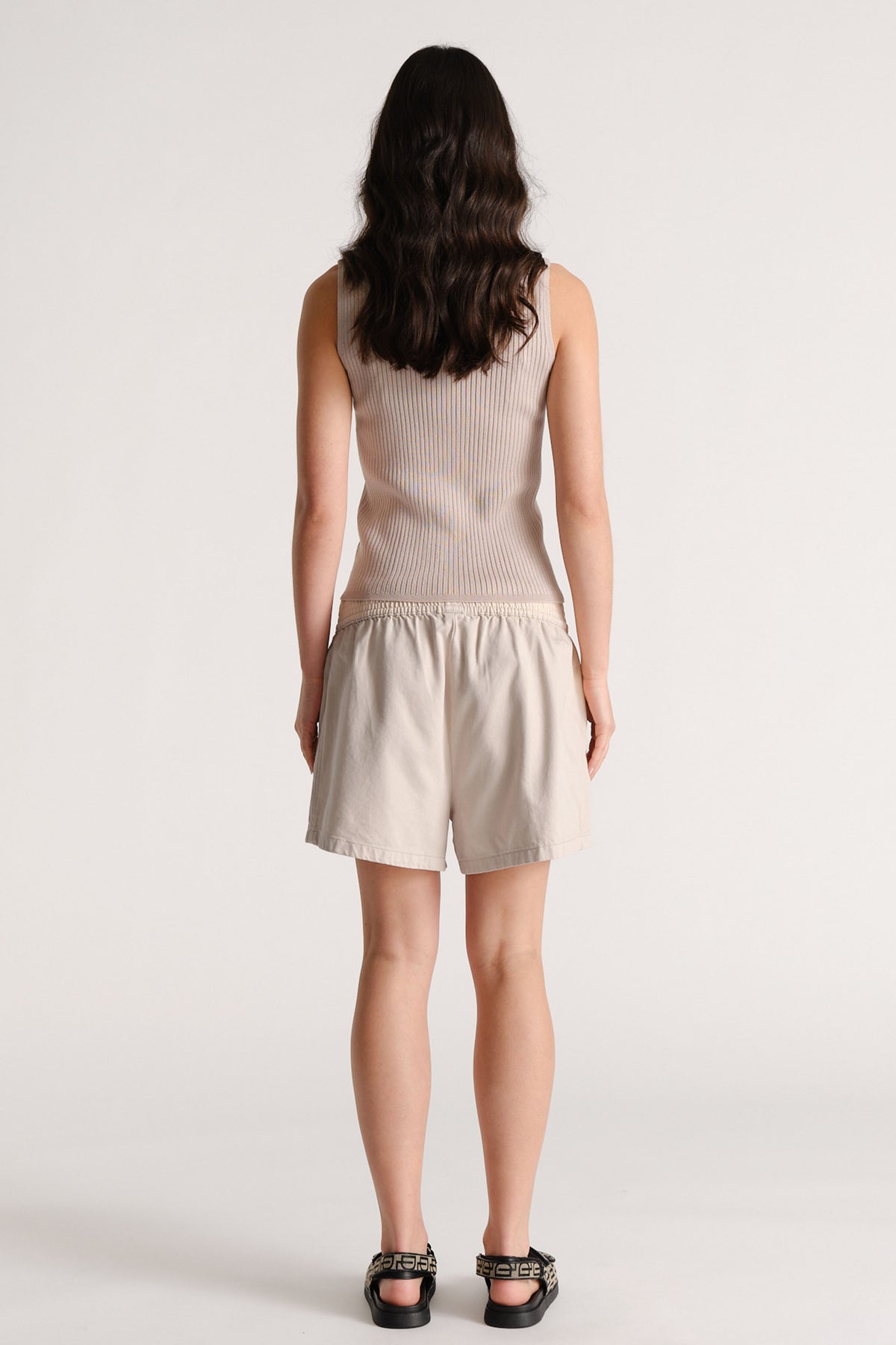 Woman seen from behind wearing a shell-colored ribbed knit top with light beige elastic waist shorts and black sandals.