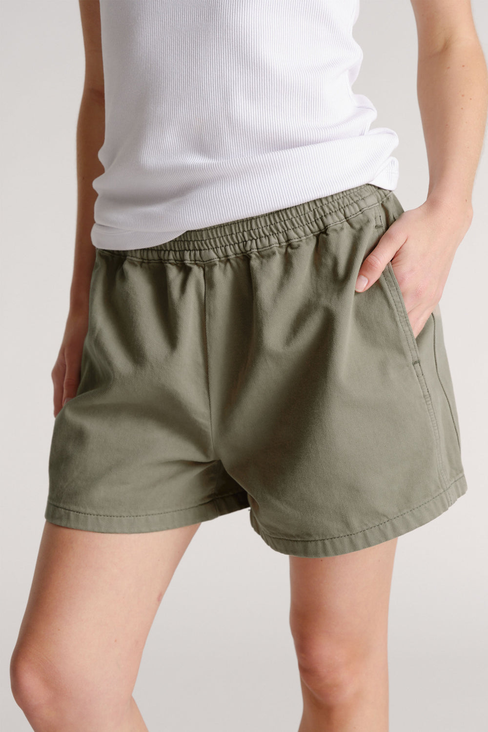Woman wearing olive green pull-on shorts with elastic waistband and side pockets paired with a white ribbed tank top