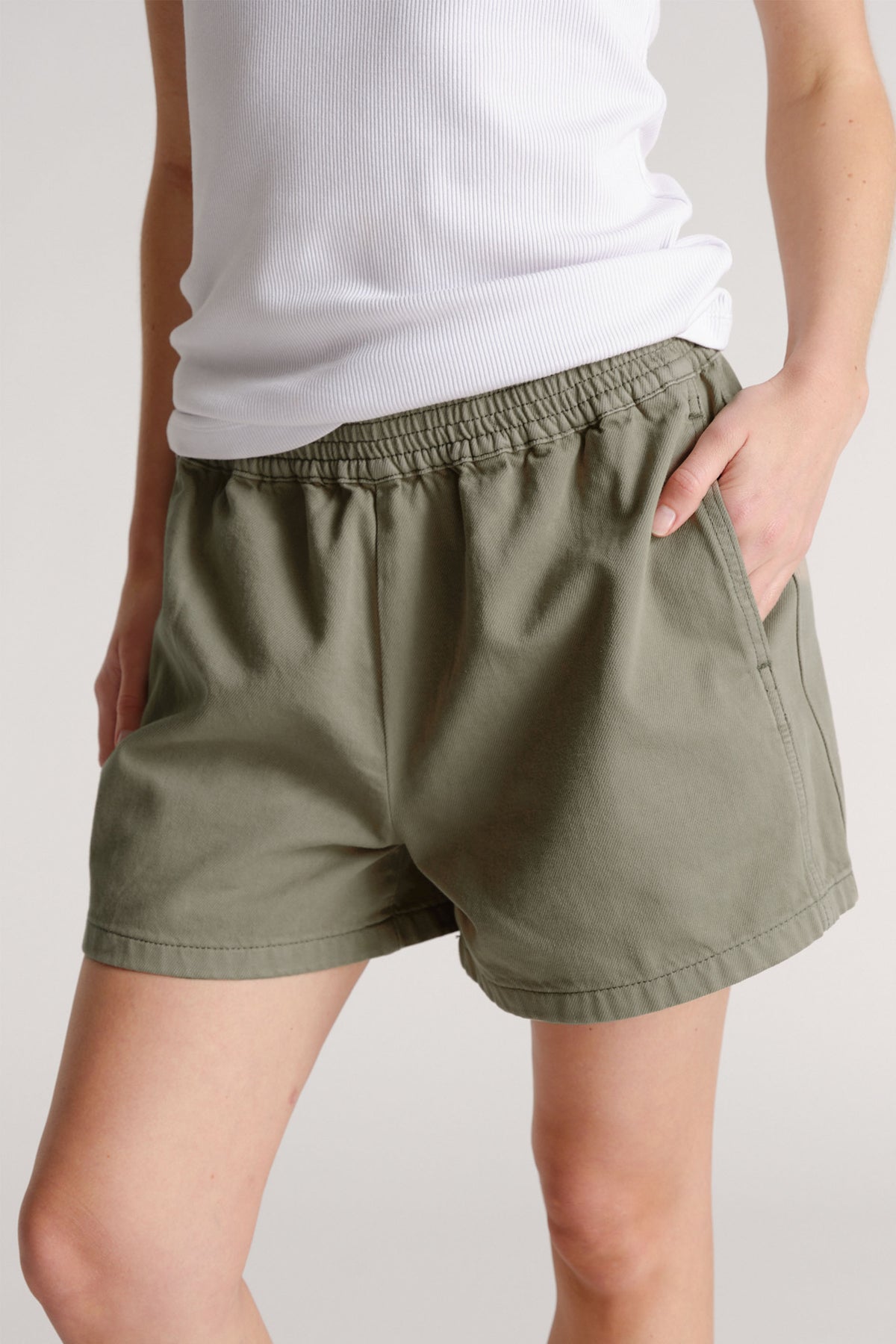 Woman wearing olive green pull-on shorts with elastic waistband and side pockets paired with a white ribbed tank top