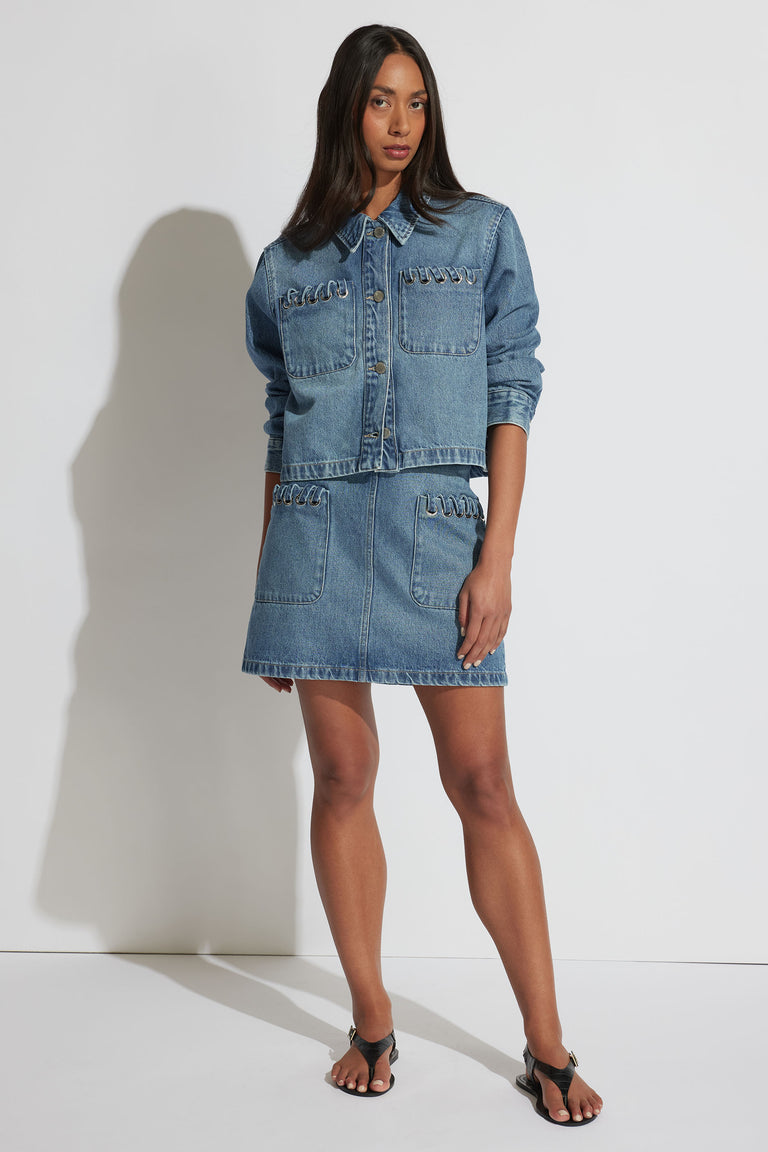 Woman wearing a bright blue denim jacket and matching skirt with unique metal ring details styled with black sandals
