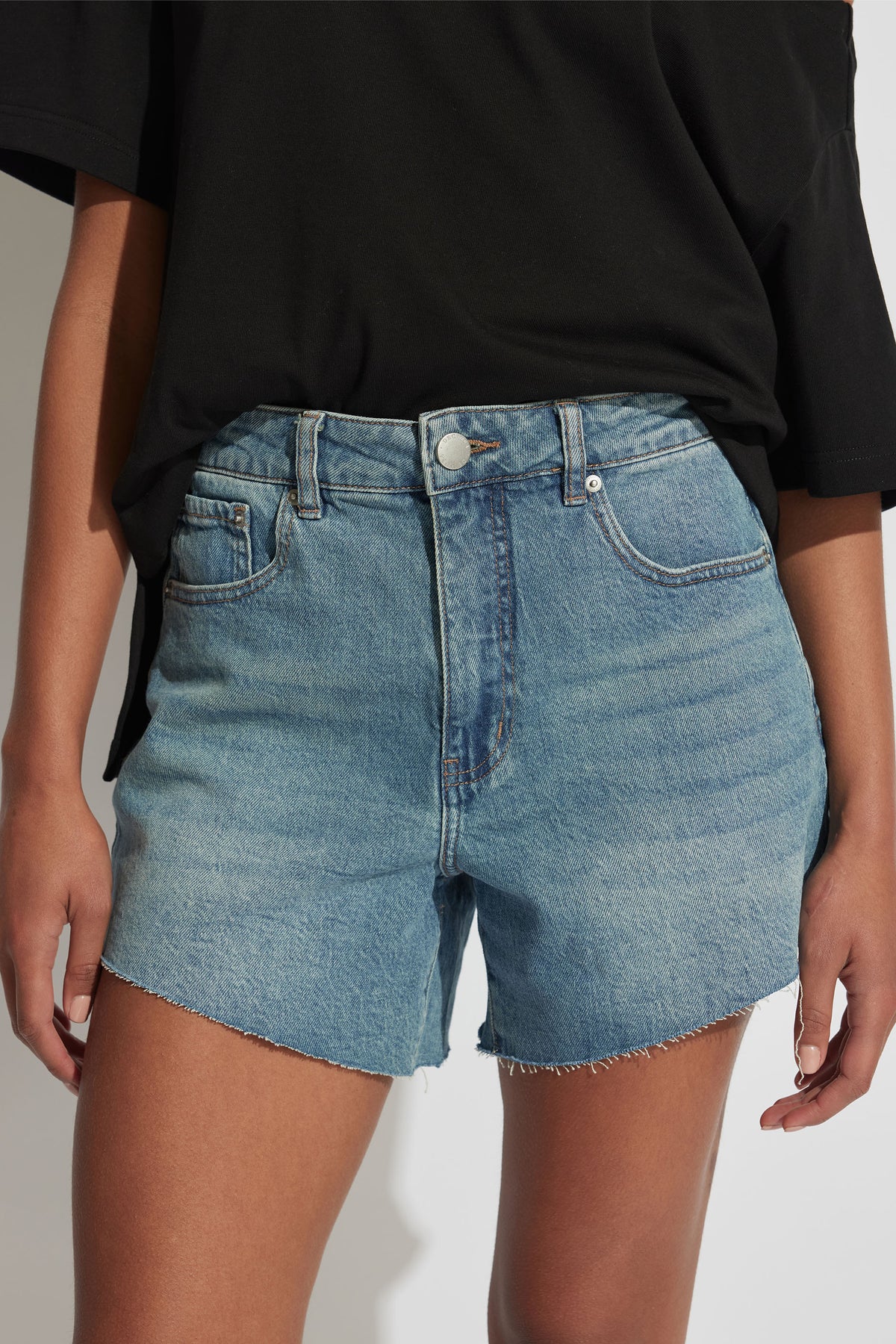 Woman wearing faded blue high-rise denim shorts with raw hem paired with a loose black tee, cropped view.