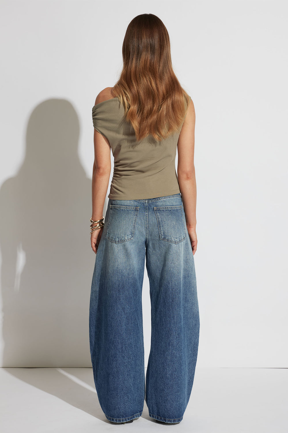 Woman wearing an olive green asymmetrical shoulder top paired with wide-leg faded blue denim jeans from the back view.