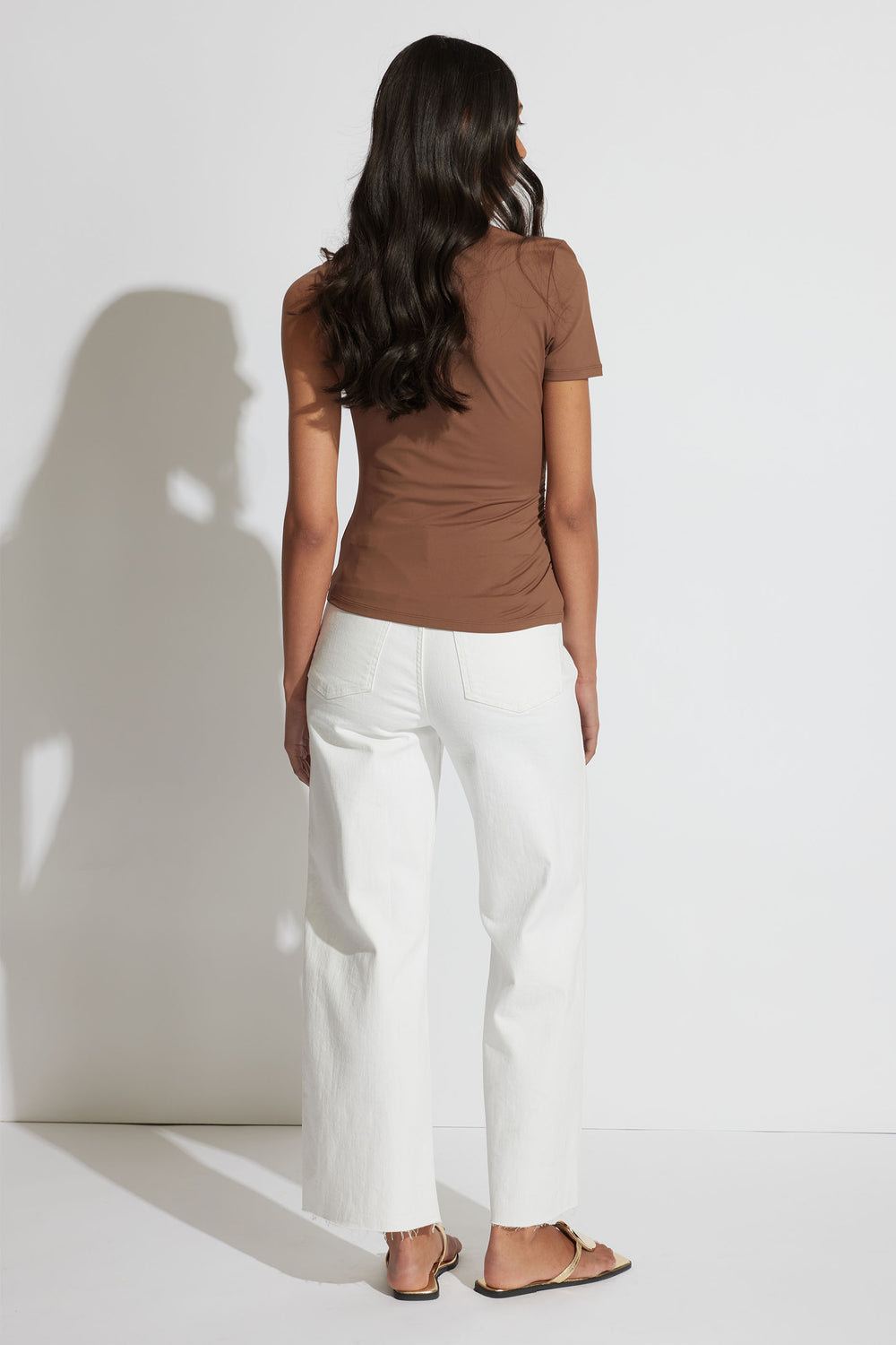 Woman wearing a fitted brown short-sleeve top paired with wide-leg white jeans and flat sandals against a plain backdrop