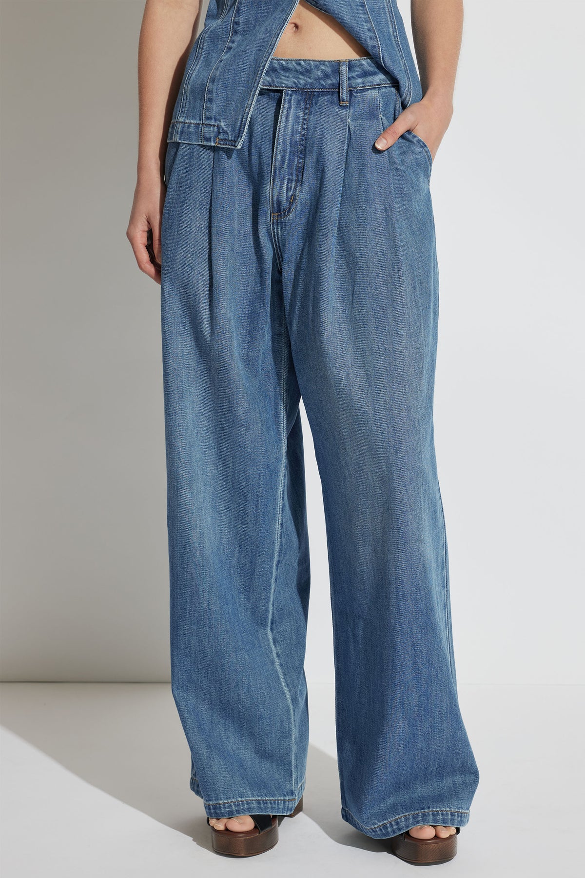 Wide-leg blue denim jeans with pleated front detail styled with open-toe brown sandals and a matching denim jacket
