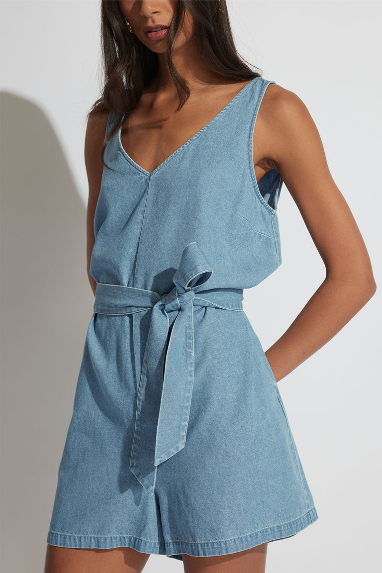 Woman wearing a light blue denim sleeveless playsuit with a V-neck and tied waist belt for a relaxed summer look