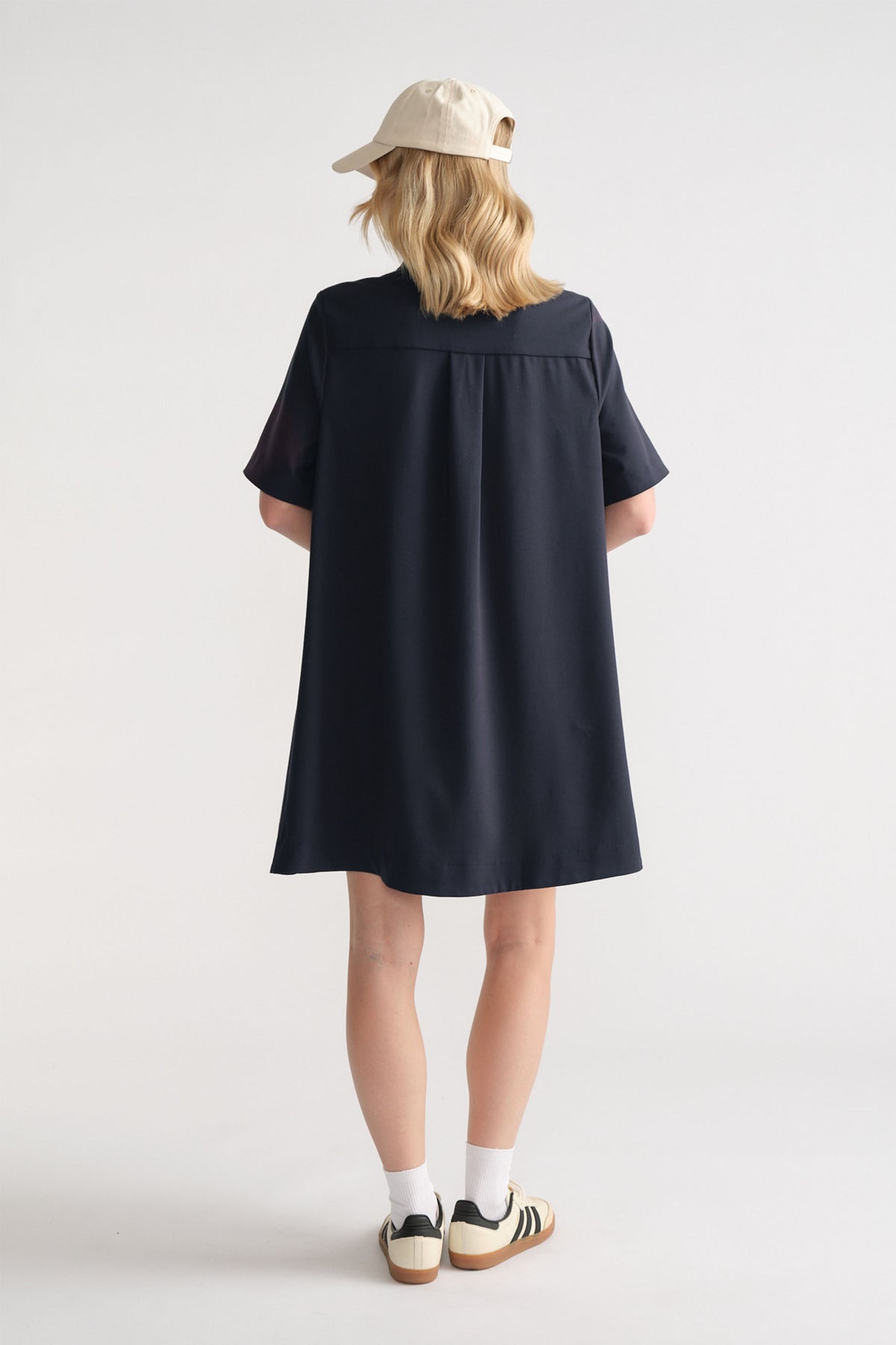 Back view of woman wearing a relaxed navy swing dress paired with white socks, sneakers, and a beige cap