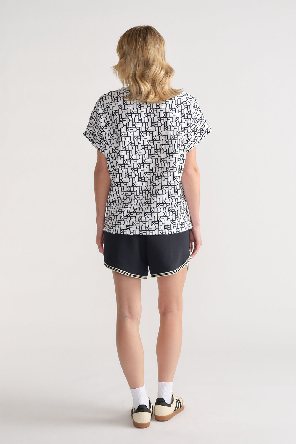 Back view of woman wearing a relaxed fit white tee with black monogram print paired with black sporty shorts.