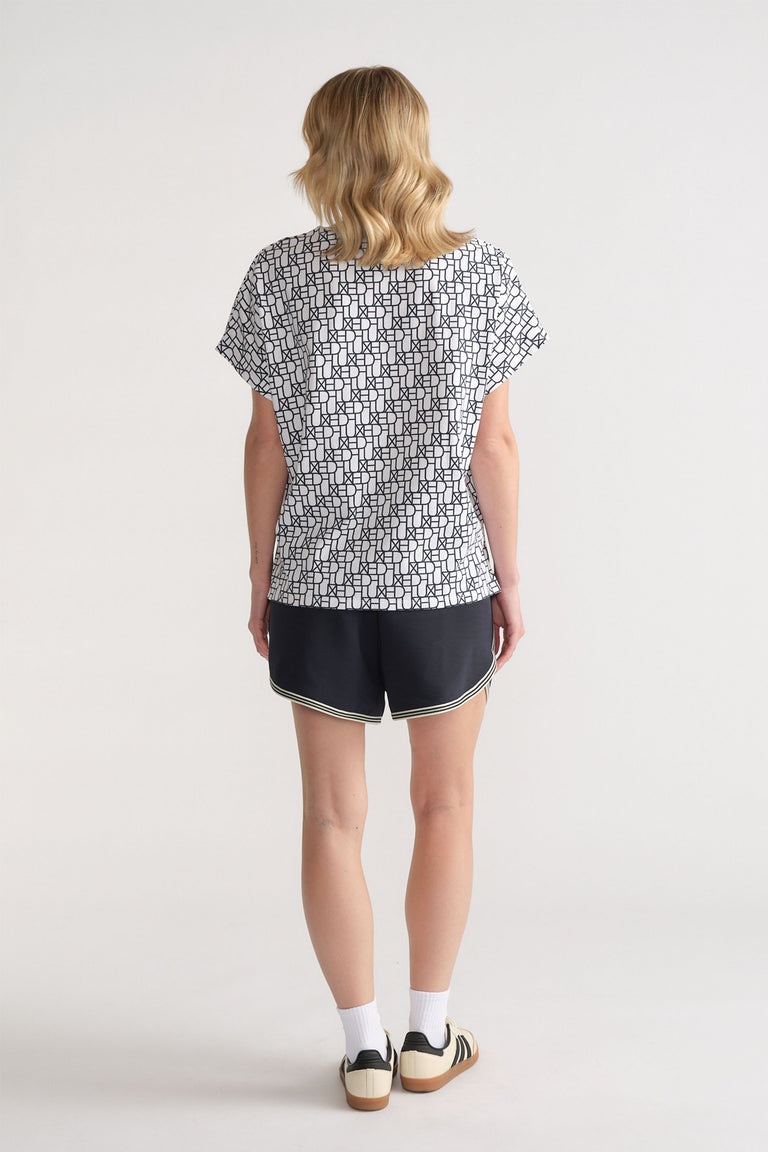 Back view of woman wearing a relaxed fit white tee with black monogram print paired with black sporty shorts.