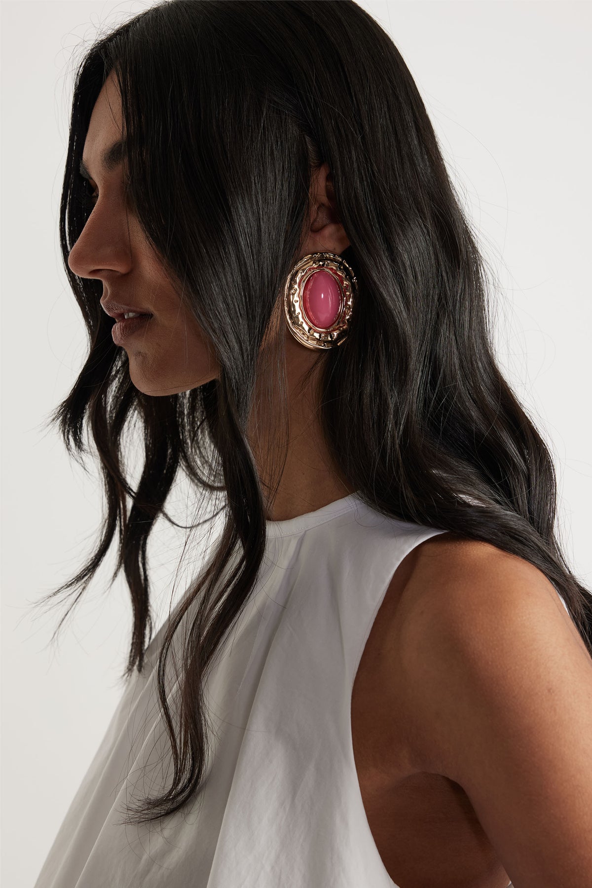 Profile view of woman wearing large gold-tone earrings with vibrant pink stones, paired with a sleeveless white top