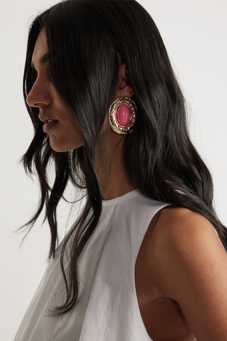 Profile view of woman wearing large gold-tone earrings with vibrant pink stones, paired with a sleeveless white top