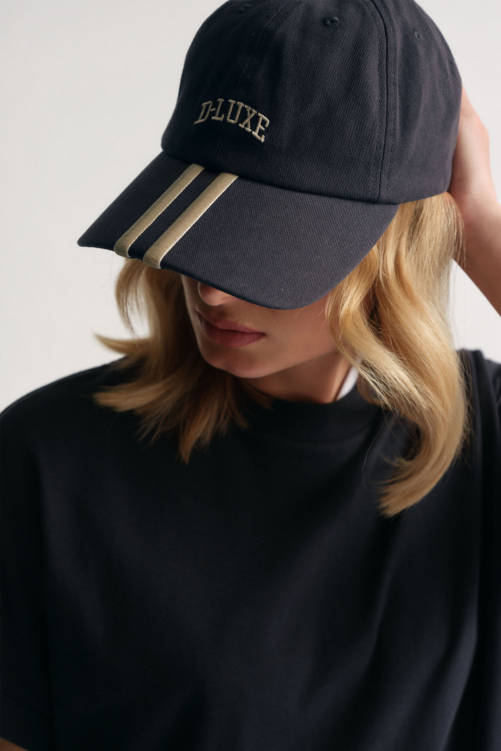 Close-up of woman wearing a navy cap with cream stripes and embroidered logo, paired with a black crewneck top.
