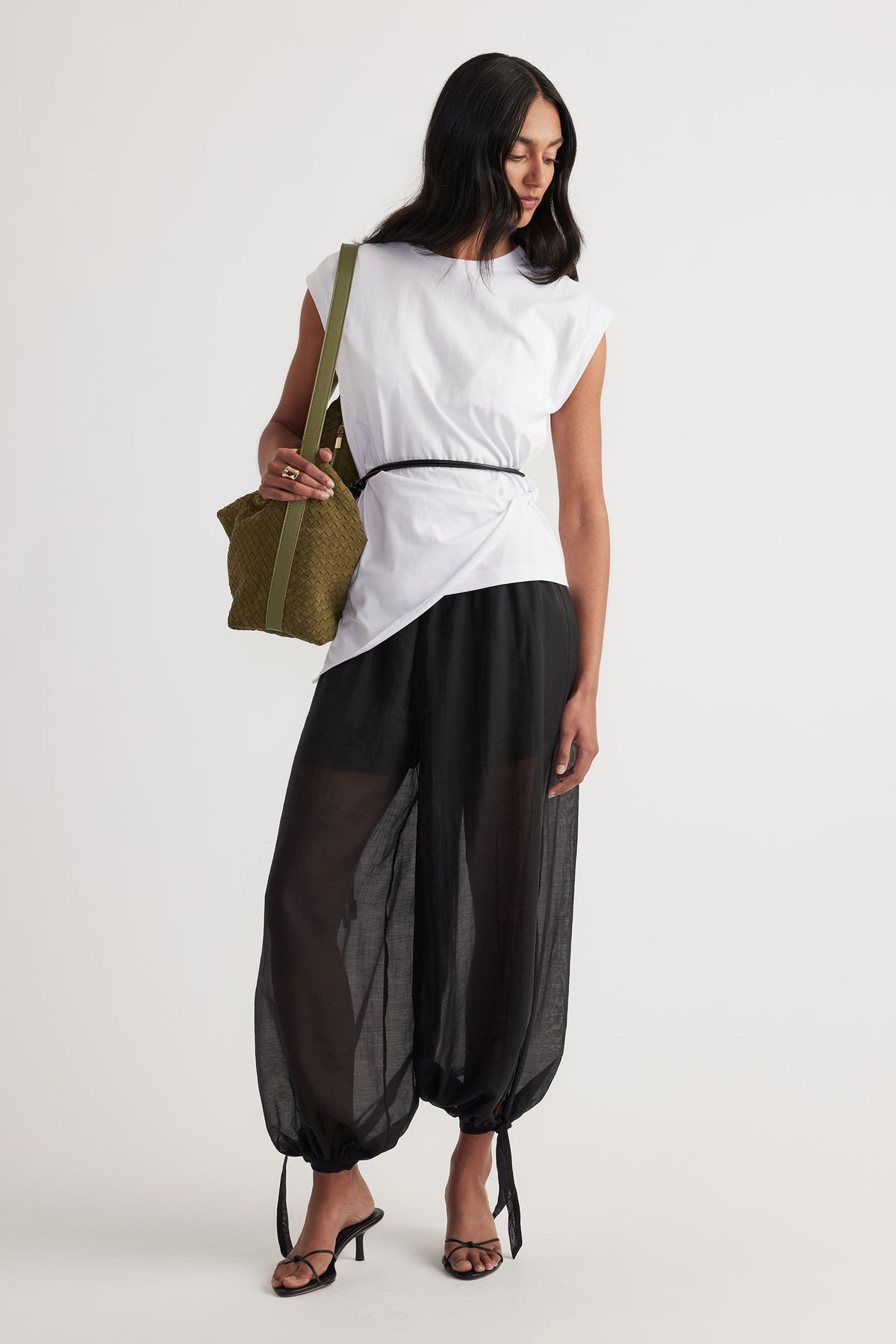 Woman wearing a white twist detail top with relaxed black sheer pants and strappy black heels, holding an olive green bag.