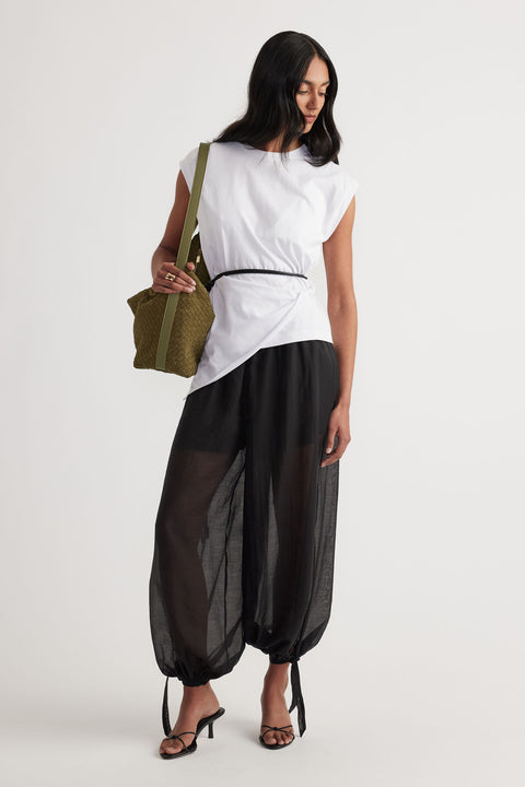 Woman wearing a white twist detail top with relaxed black sheer pants and strappy black heels, holding an olive green bag.