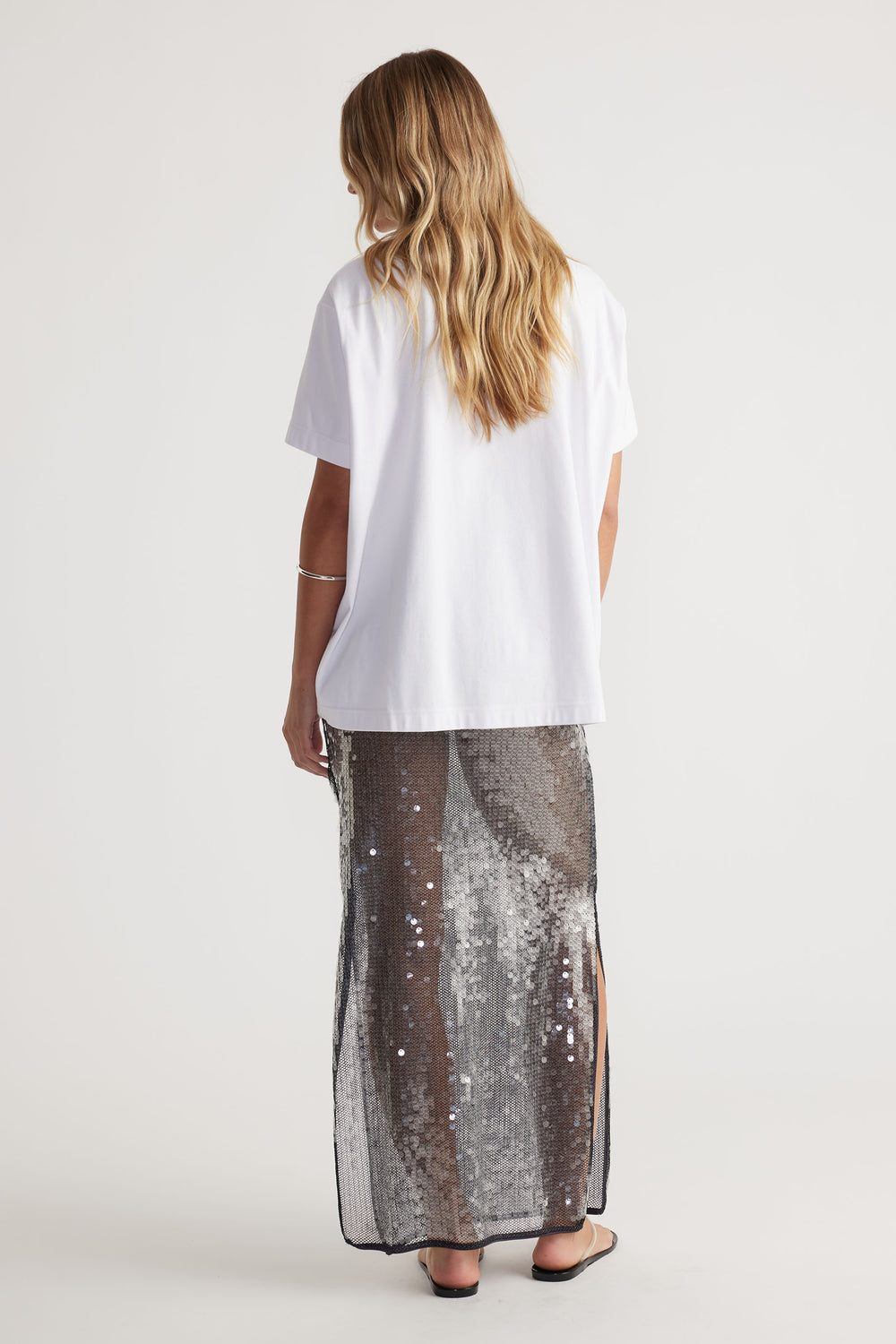 Woman wearing a white t-shirt and Savannah Sequin Skirt with side splits, elastic waist, and semi sheer maxi length back view.