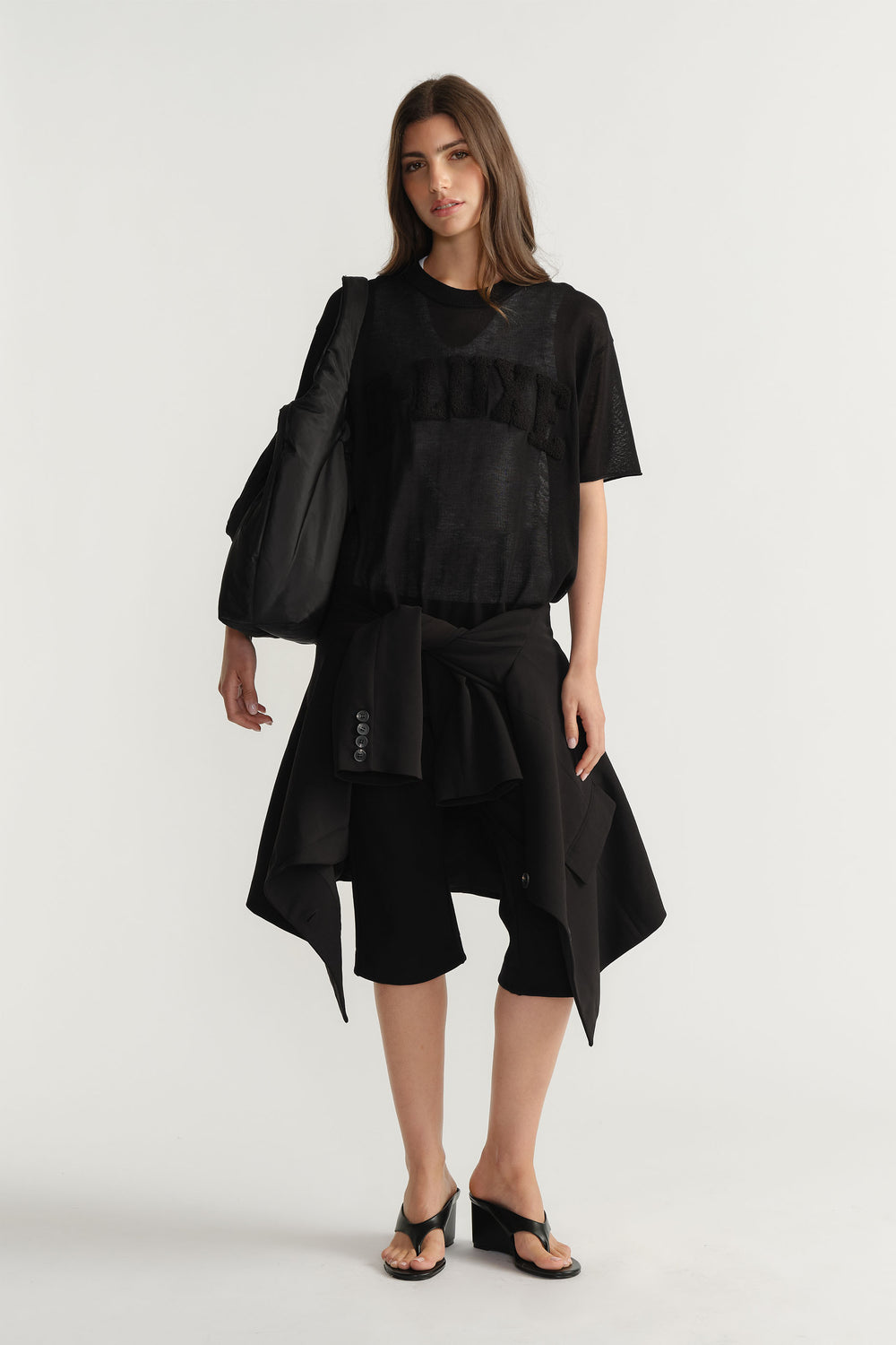 Portrait of model wearing black D-Luxe Fine Knit Tee with semi-sheer bouclé logo, styled with black shorts and jacket, gallery 1.
