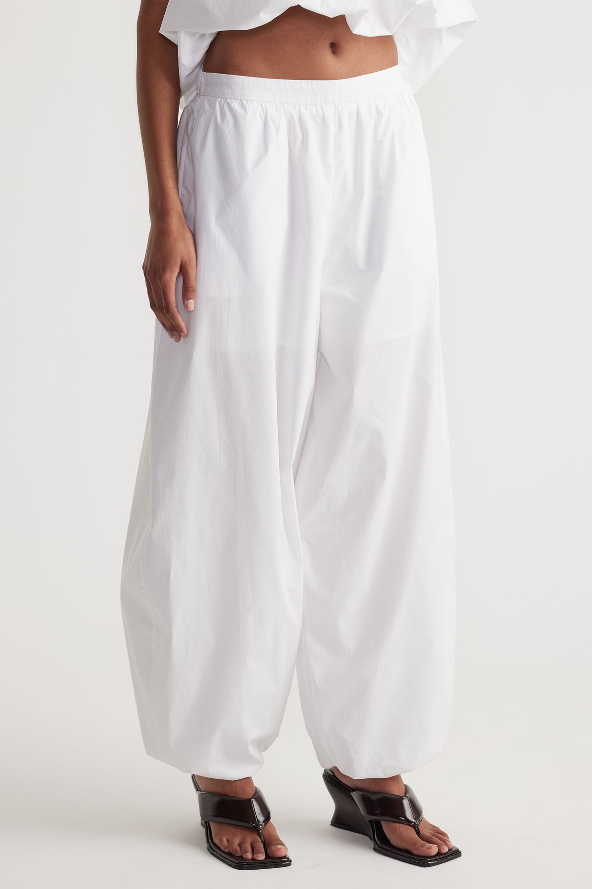 Front view of white cotton balloon pants with elastic waist, paired with black open-toe wedge sandals.