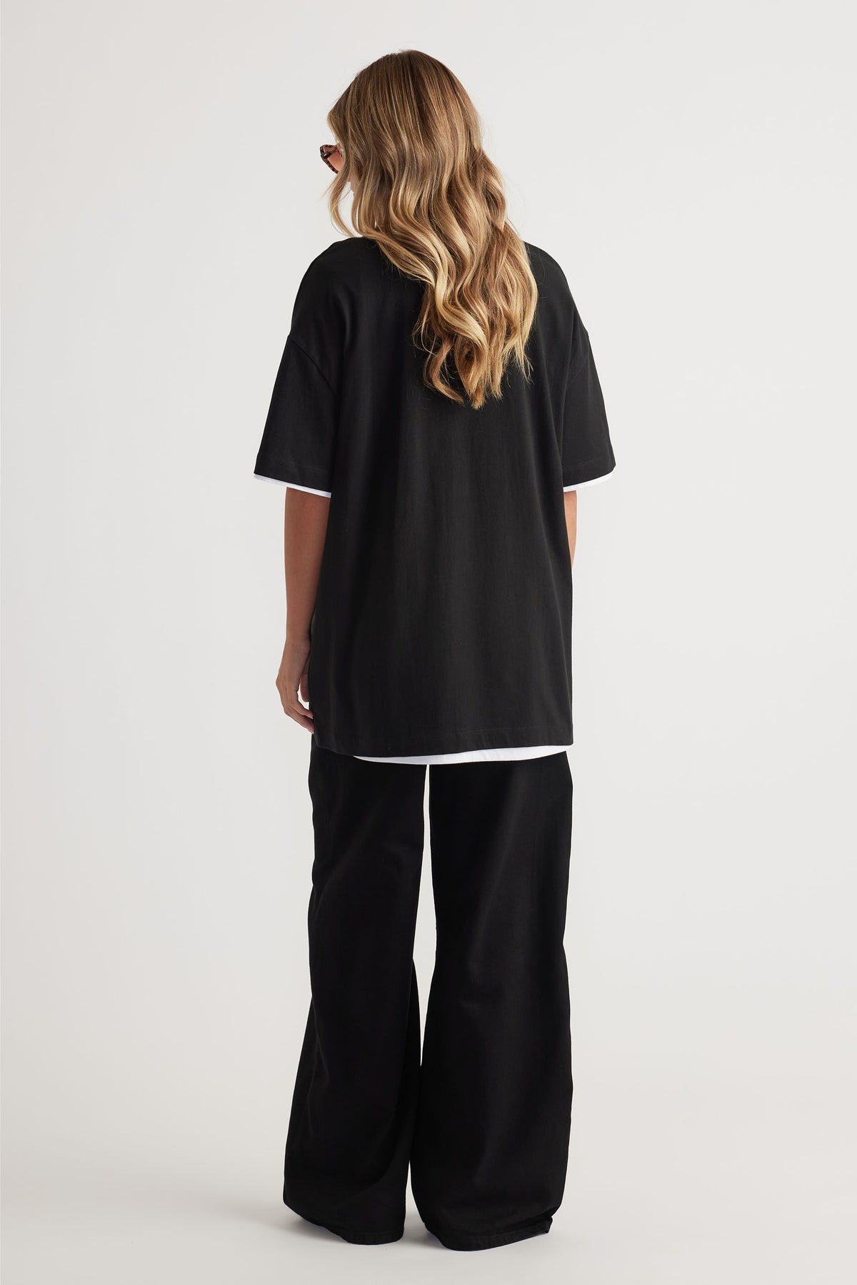 Rear view of model wearing black Kayla Longline Tee layered over white tee with wide-leg black pants by Decjuba Australian brand