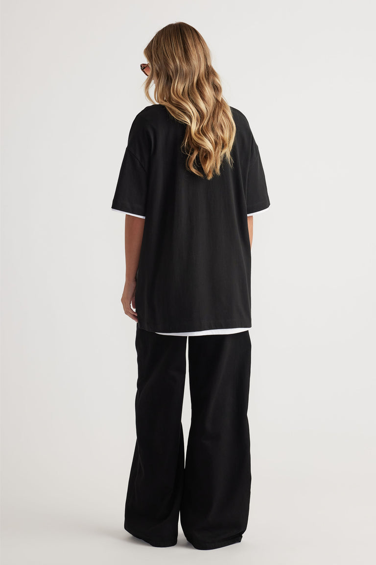 Rear view of model wearing black Kayla Longline Tee layered over white tee with wide-leg black pants by Decjuba Australian brand