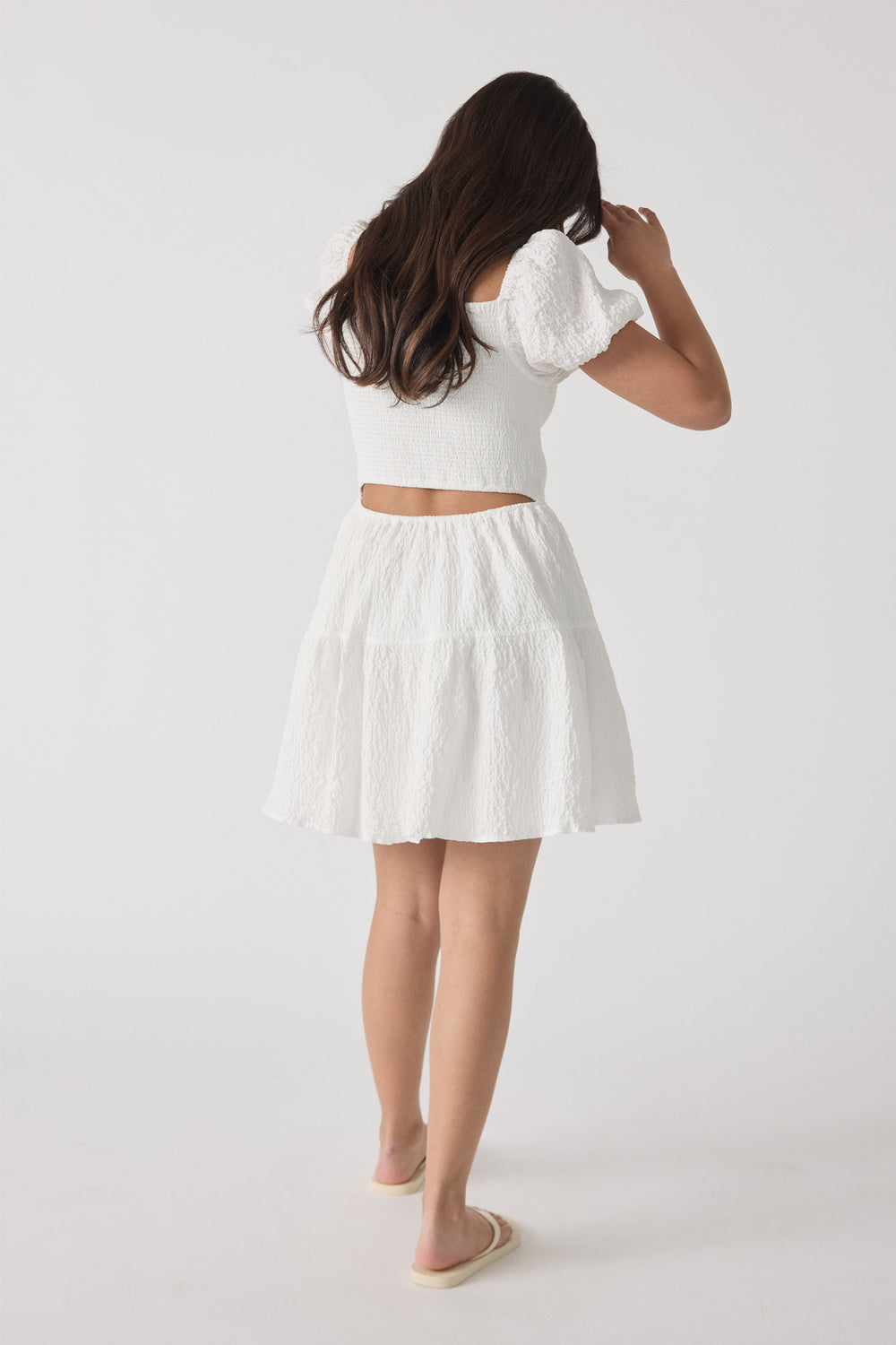 Girl wearing a textured white mini dress with puff sleeves and a cutout back, paired with slip-on sandals.