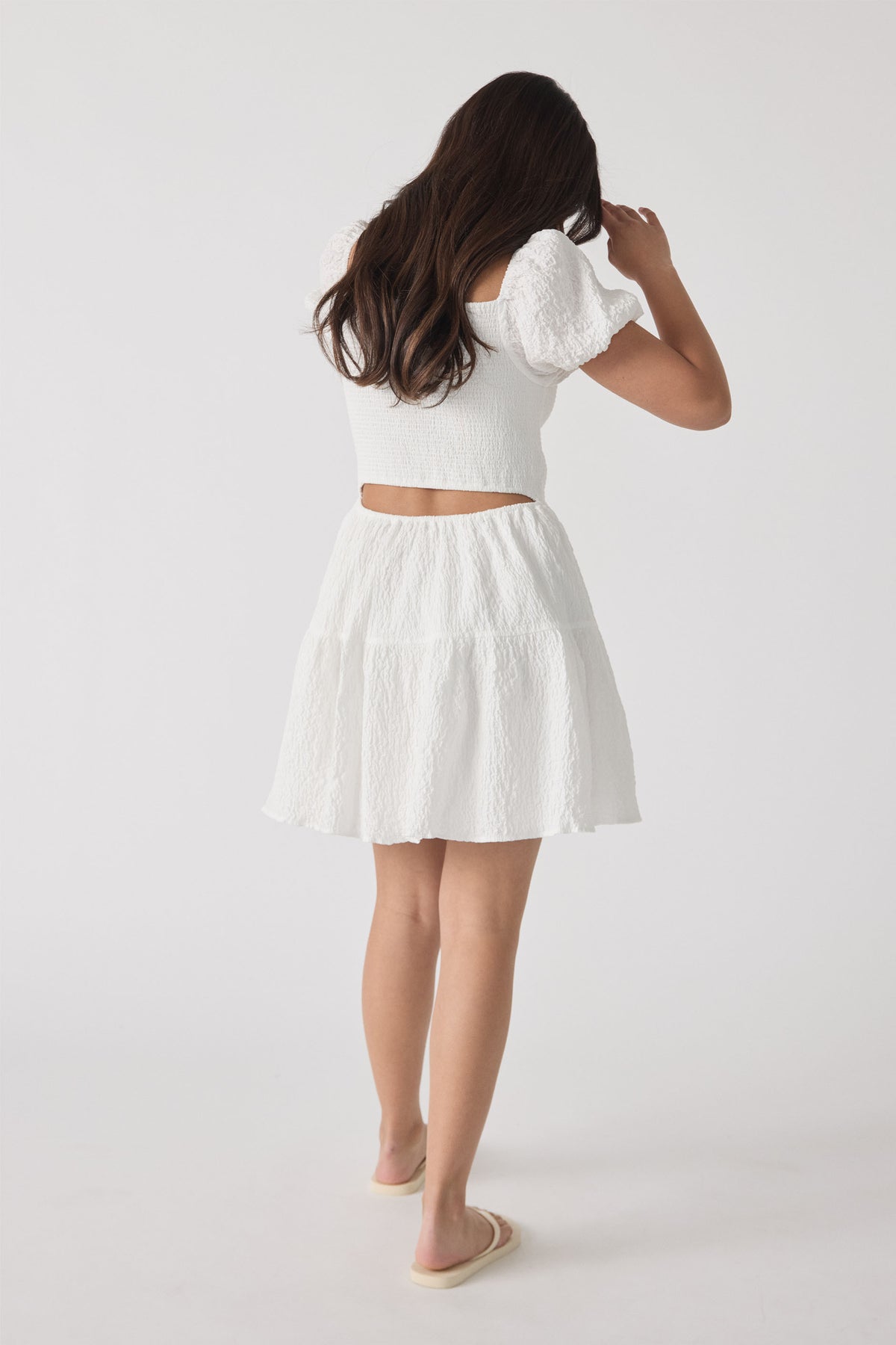 Girl wearing a textured white mini dress with puff sleeves and a cutout back, paired with slip-on sandals.
