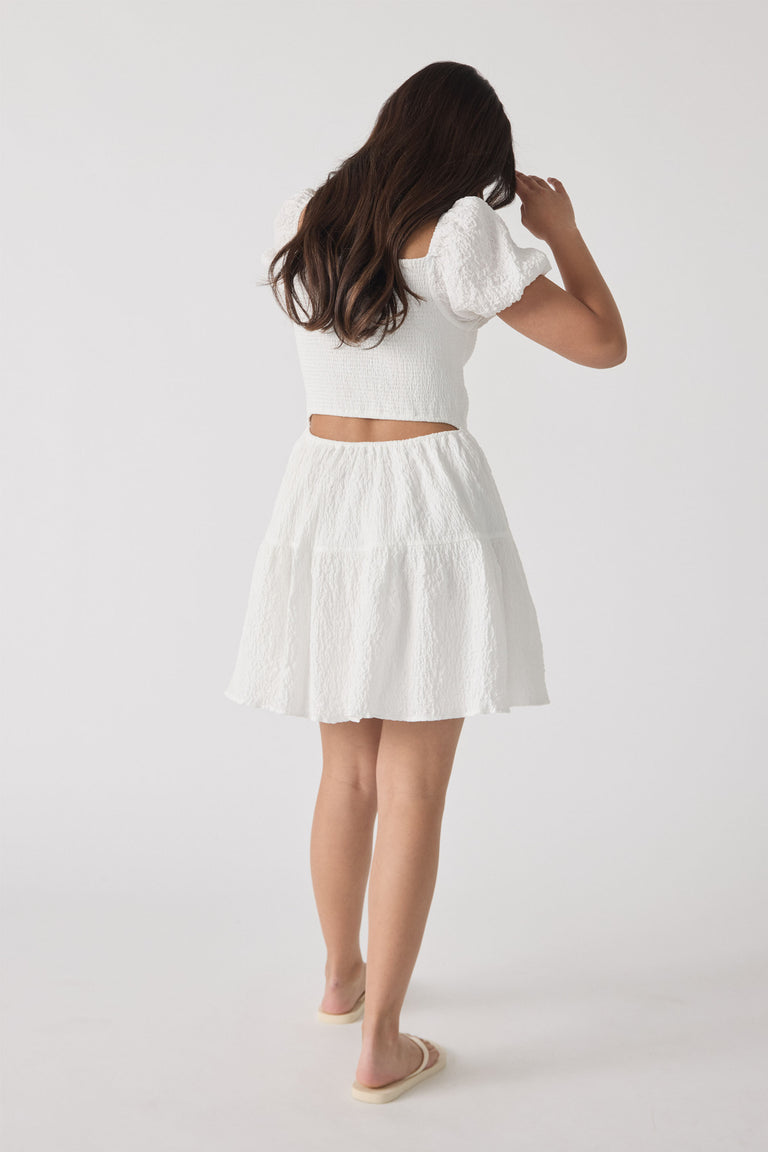 Girl wearing a textured white mini dress with puff sleeves and a cutout back, paired with slip-on sandals.