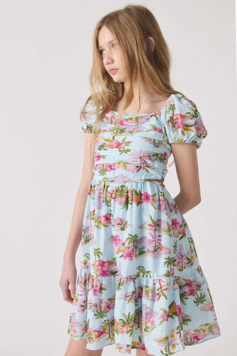 Girl wearing a light blue mini dress with pink floral and palm print, featuring puff sleeves and a gathered waist.
