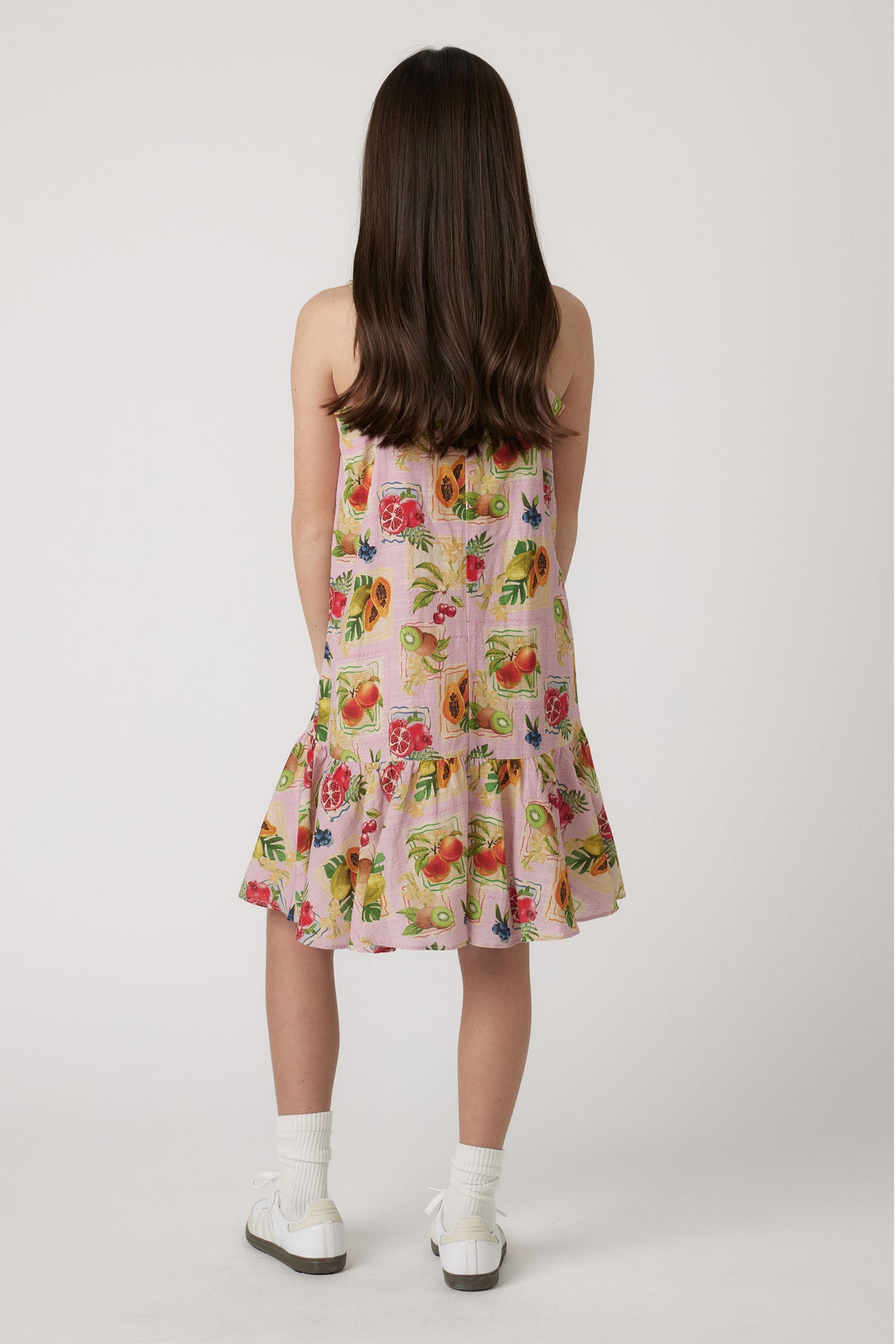 Girl wearing a pink strappy dress with colorful fruit print, paired with white socks and sneakers seen from behind