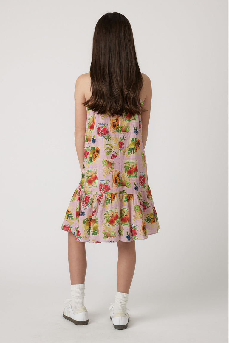 Girl wearing a pink strappy dress with colorful fruit print, paired with white socks and sneakers seen from behind