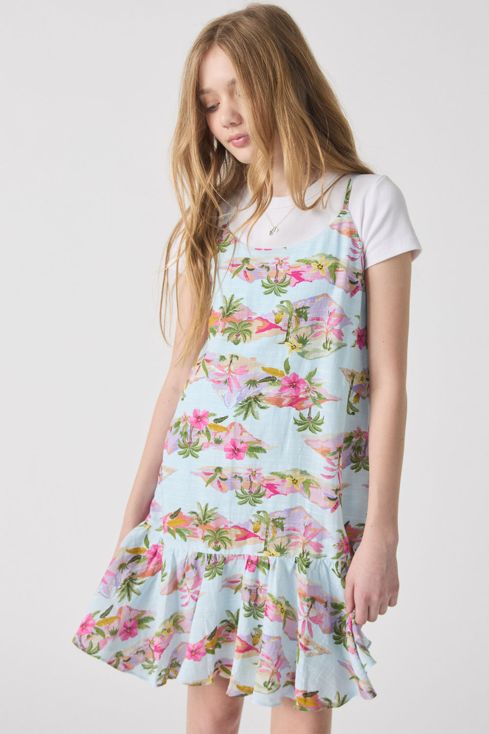 Girl wearing a light blue floral strappy dress over a white tee, featuring tropical palm and pink flower prints with a ruffled hem.