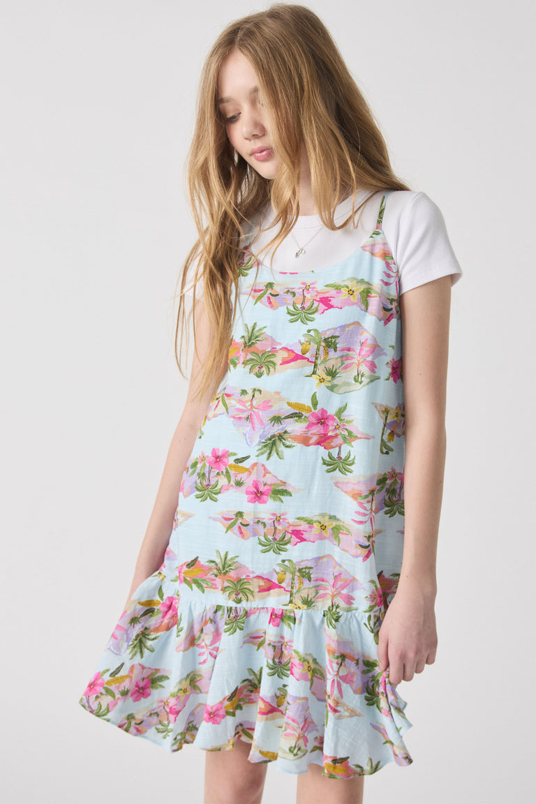 Girl wearing a light blue floral strappy dress over a white tee, featuring tropical palm and pink flower prints with a ruffled hem.