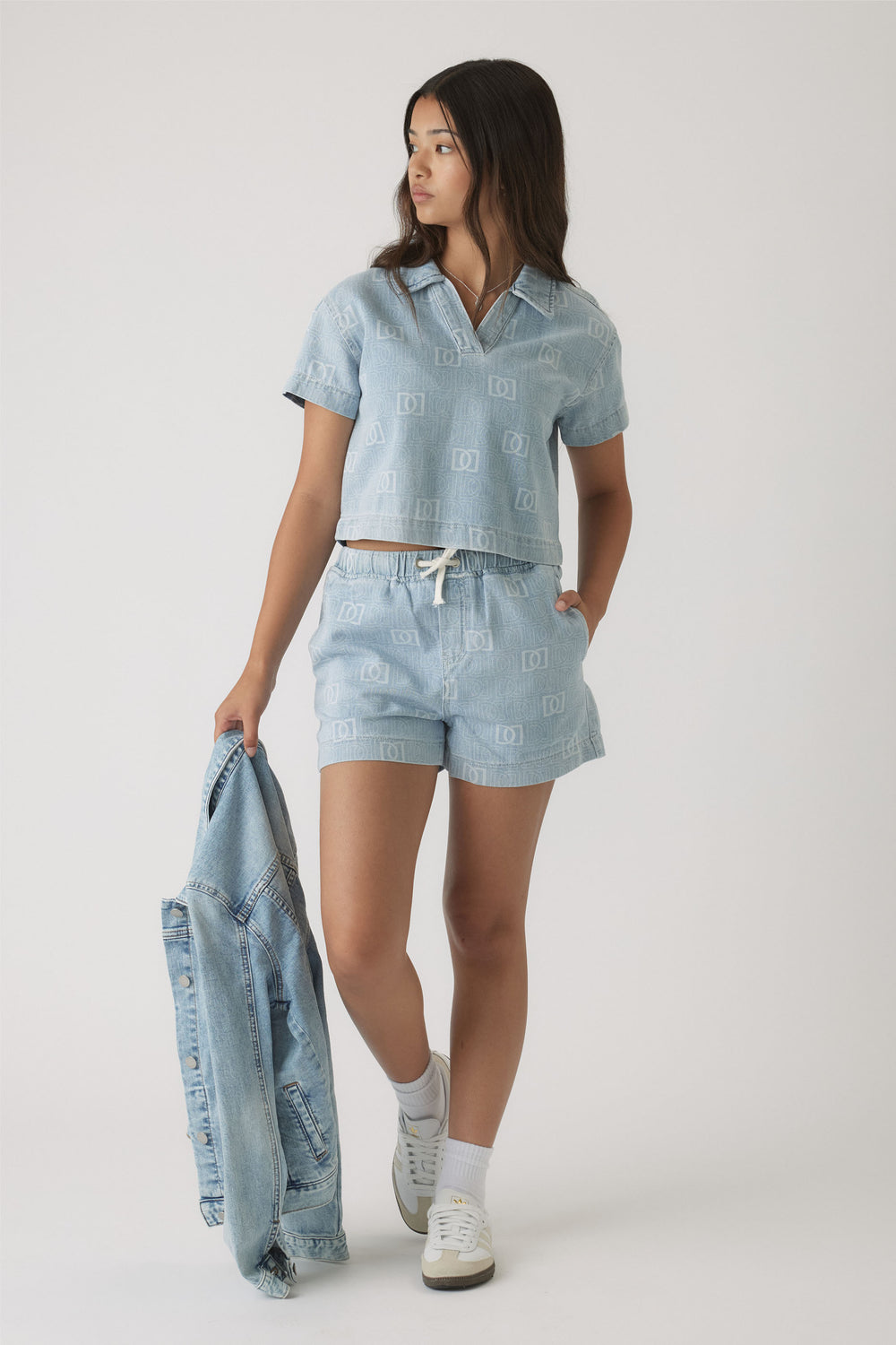 Teen girl wearing light blue denim shorts and a matching short-sleeve shirt with subtle monogram print, holding a denim jacket