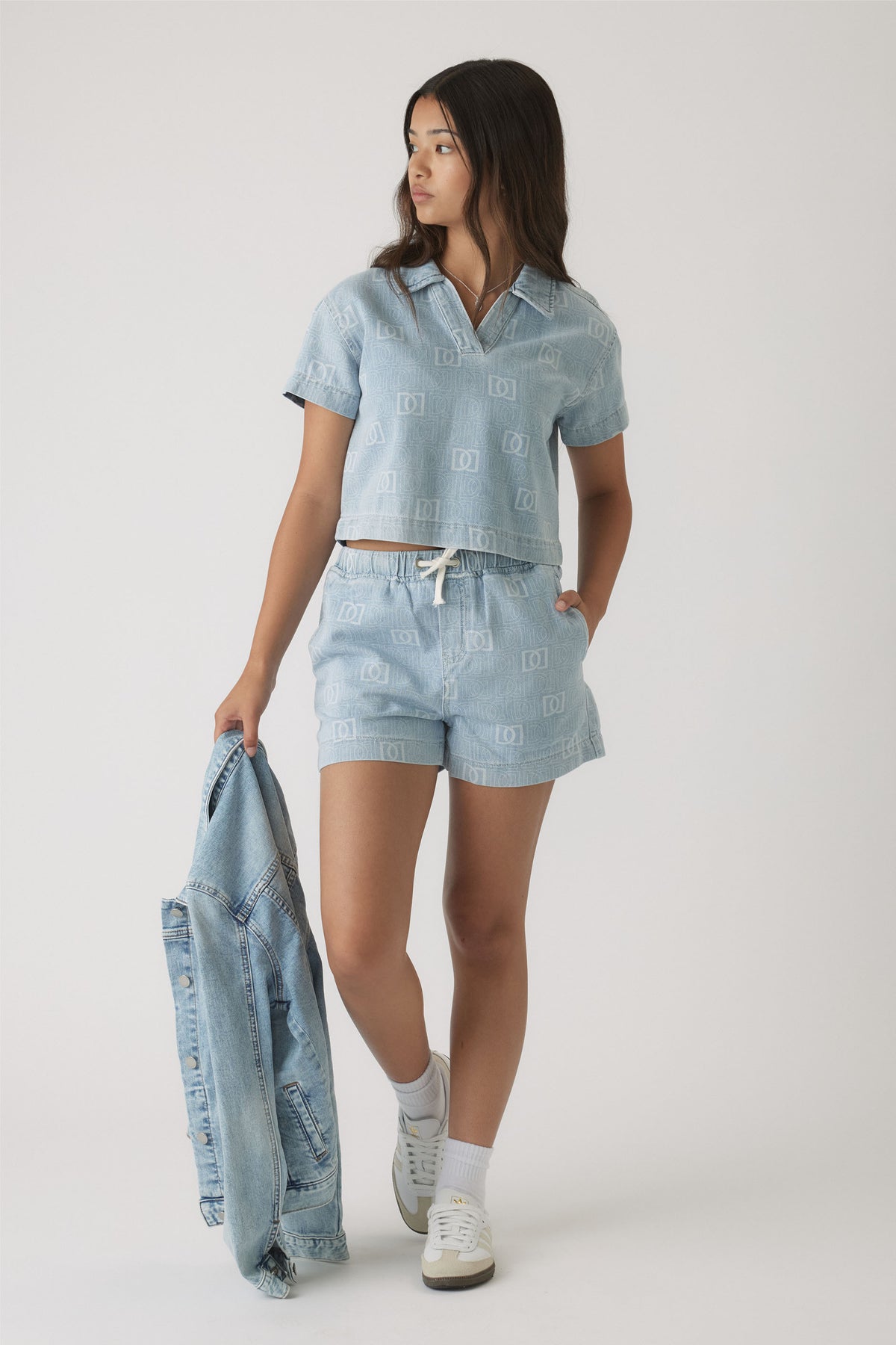 Teen girl wearing light blue denim shorts and a matching short-sleeve shirt with subtle monogram print, holding a denim jacket