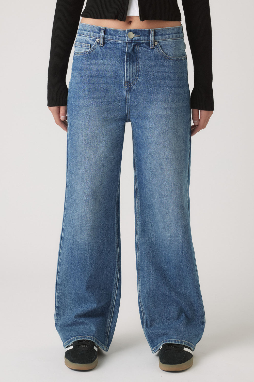 Front view of woman wearing high-rise wide-leg blue denim jeans paired with a cropped black long-sleeve top and black sneakers