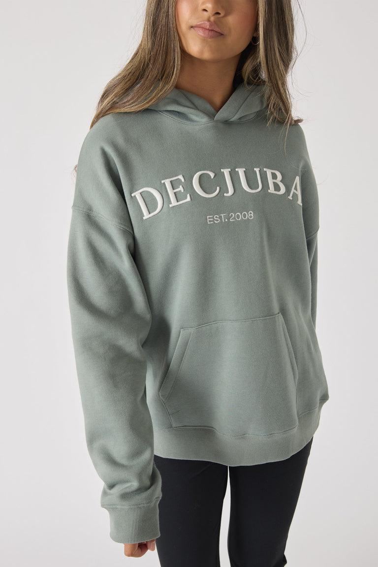 Girl wearing a soft sage green hoodie with a front kangaroo pocket and bold embroidered logo across the chest