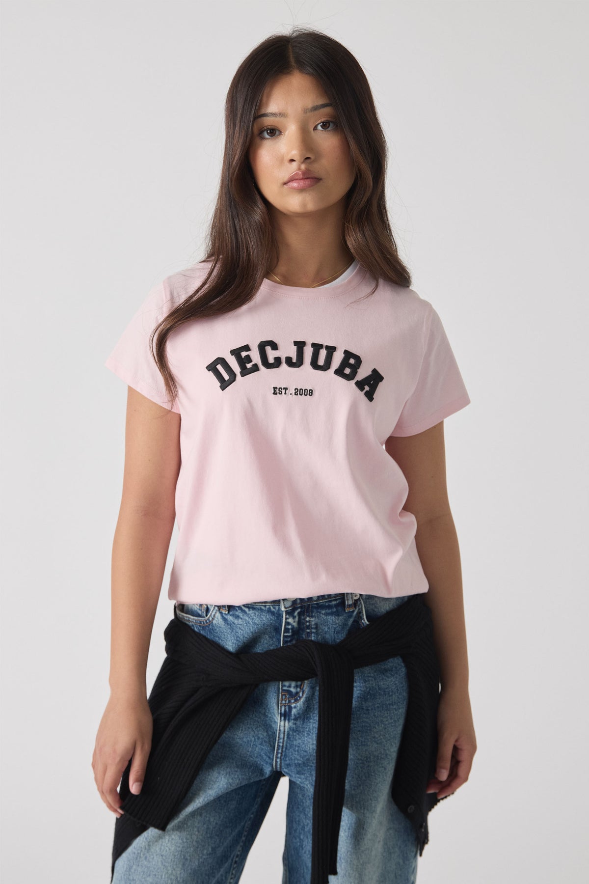 Teen girl wearing a soft pink tee with bold black lettering, styled with light wash jeans and a tied black sweater.