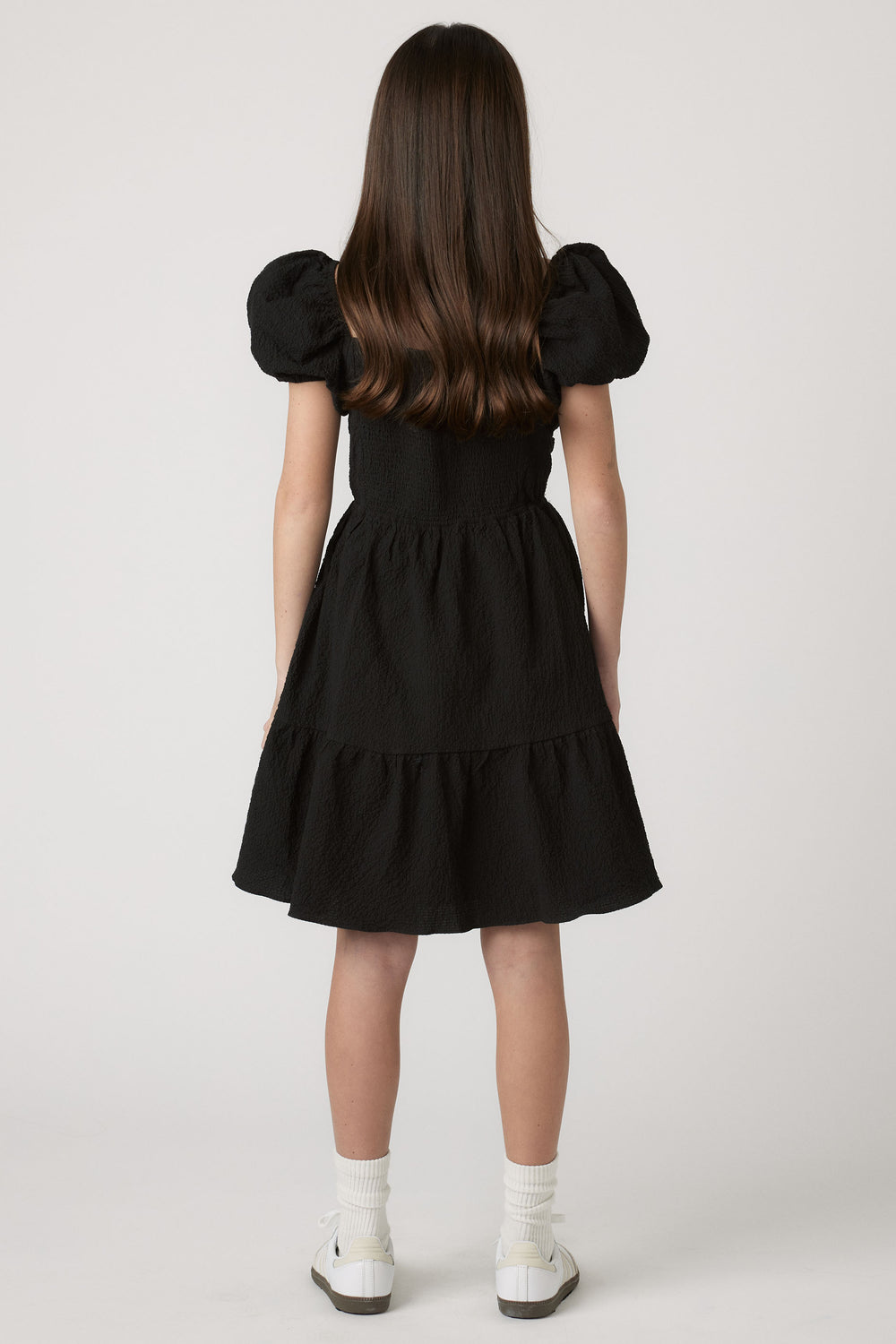 Girl wearing a black textured mini dress with puff sleeves, paired with white socks and casual sneakers for easy style.