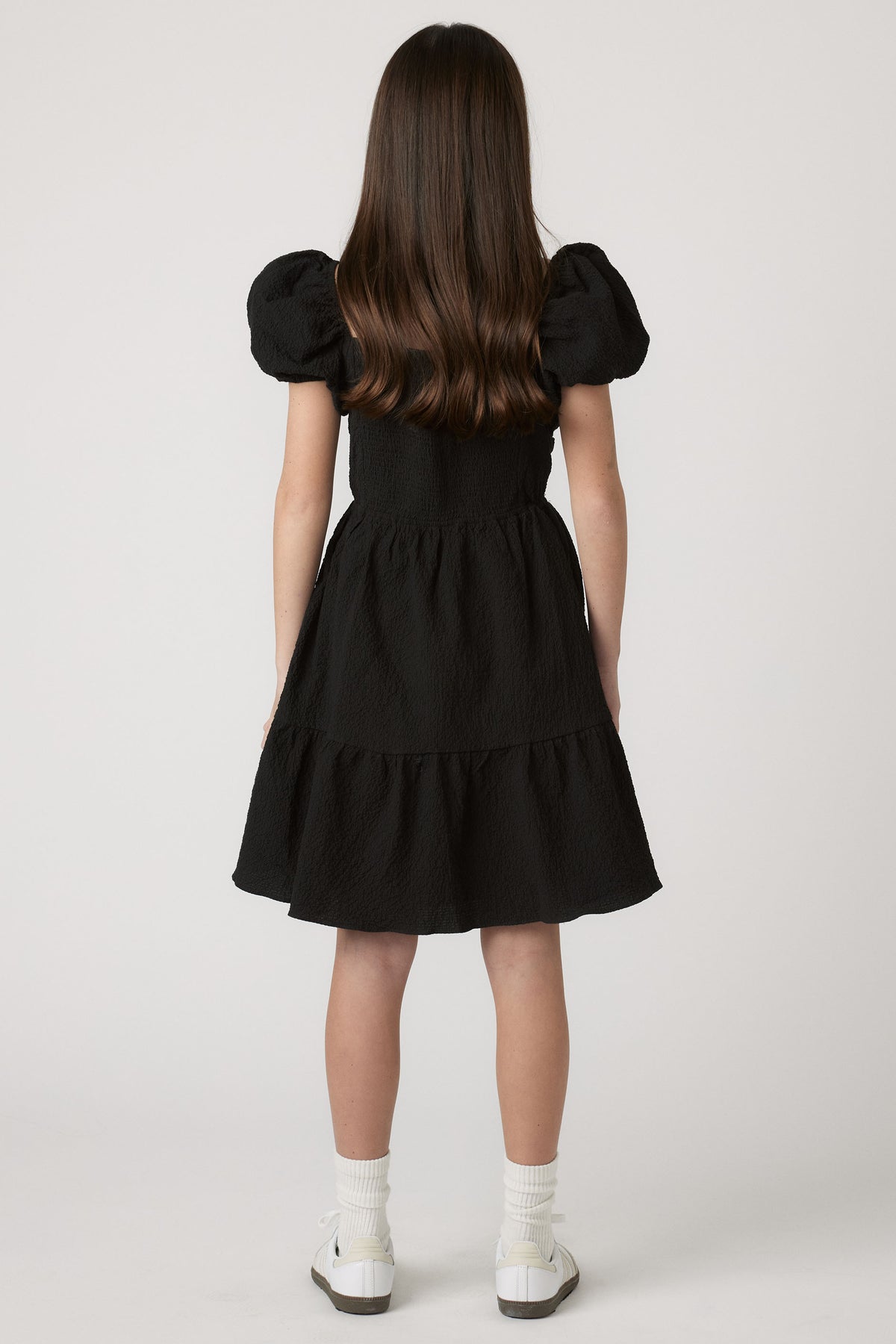 Girl wearing a black textured mini dress with puff sleeves, paired with white socks and casual sneakers for easy style.