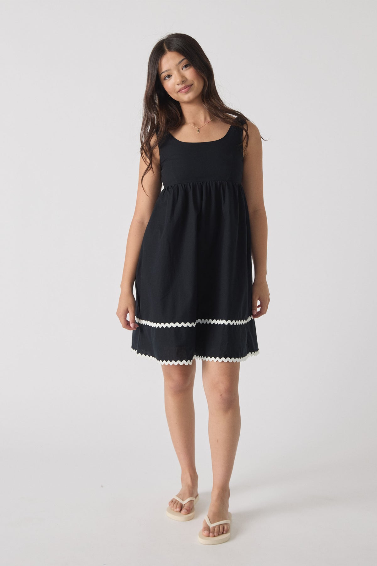 Girl wearing a sleeveless black mini dress with white trim details, styled with casual flip-flops, standing against plain background.