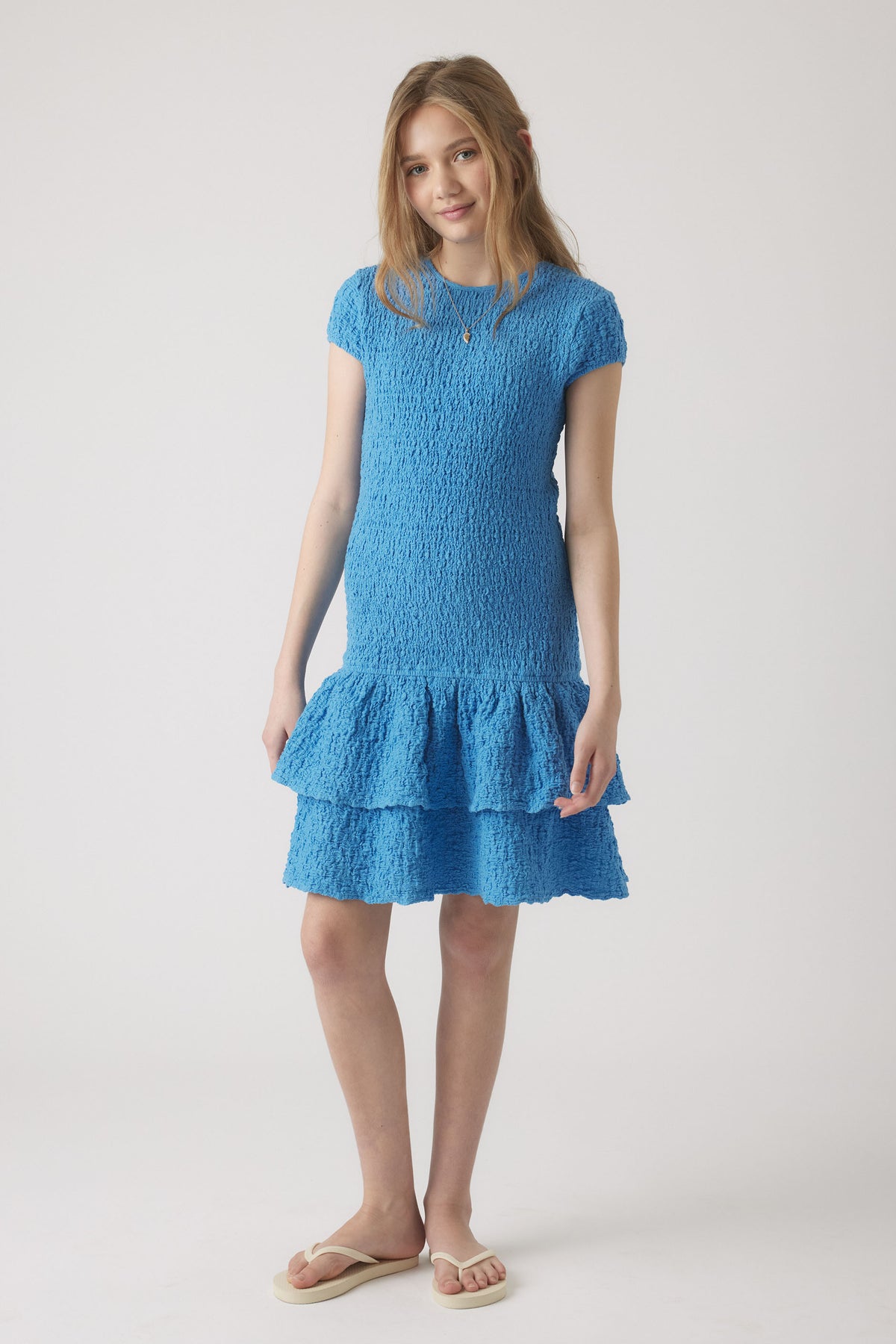 Girl wearing a textured bright blue shirred dress with short sleeves and tiered ruffle hem, styled with simple sandals.