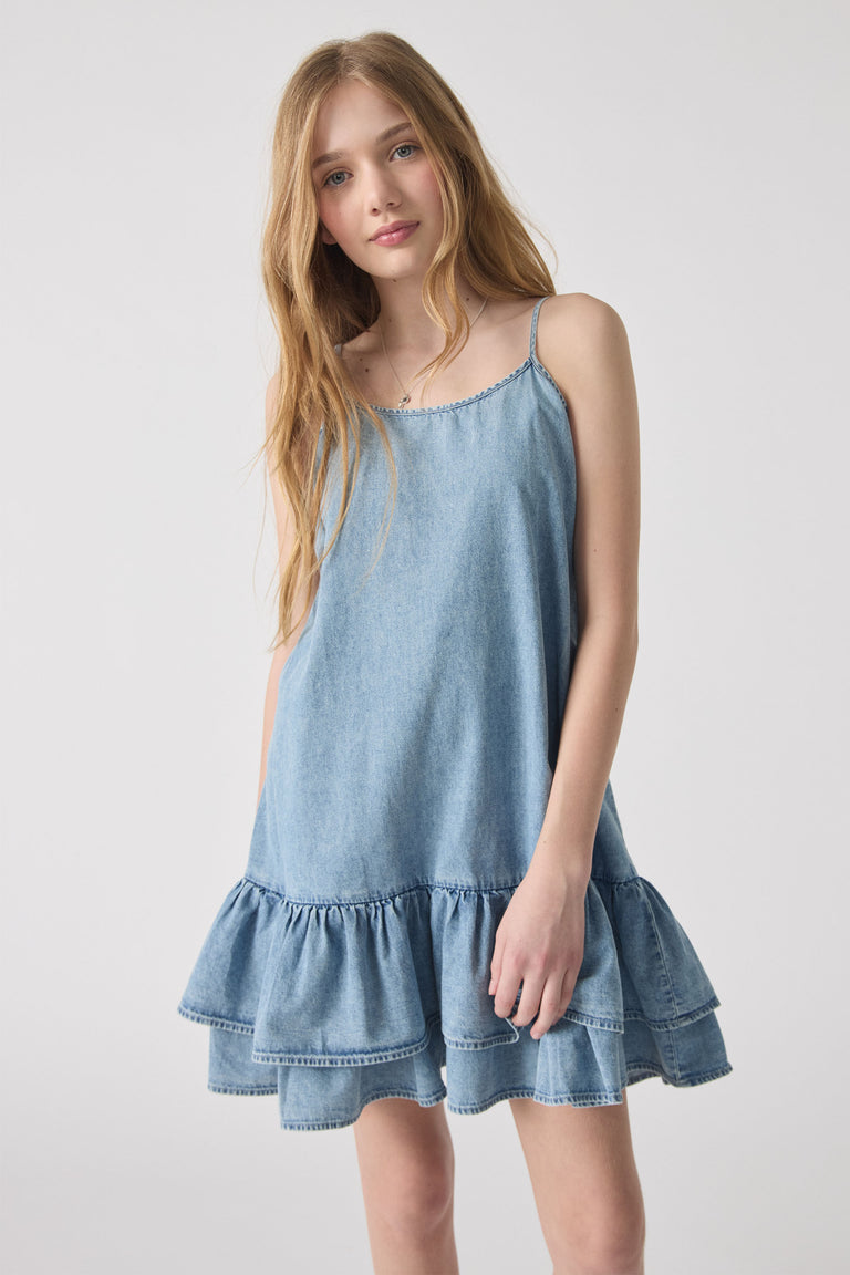 Girl wearing a light blue denim dress with thin straps and a tiered ruffle hem, styled casually for everyday wear.